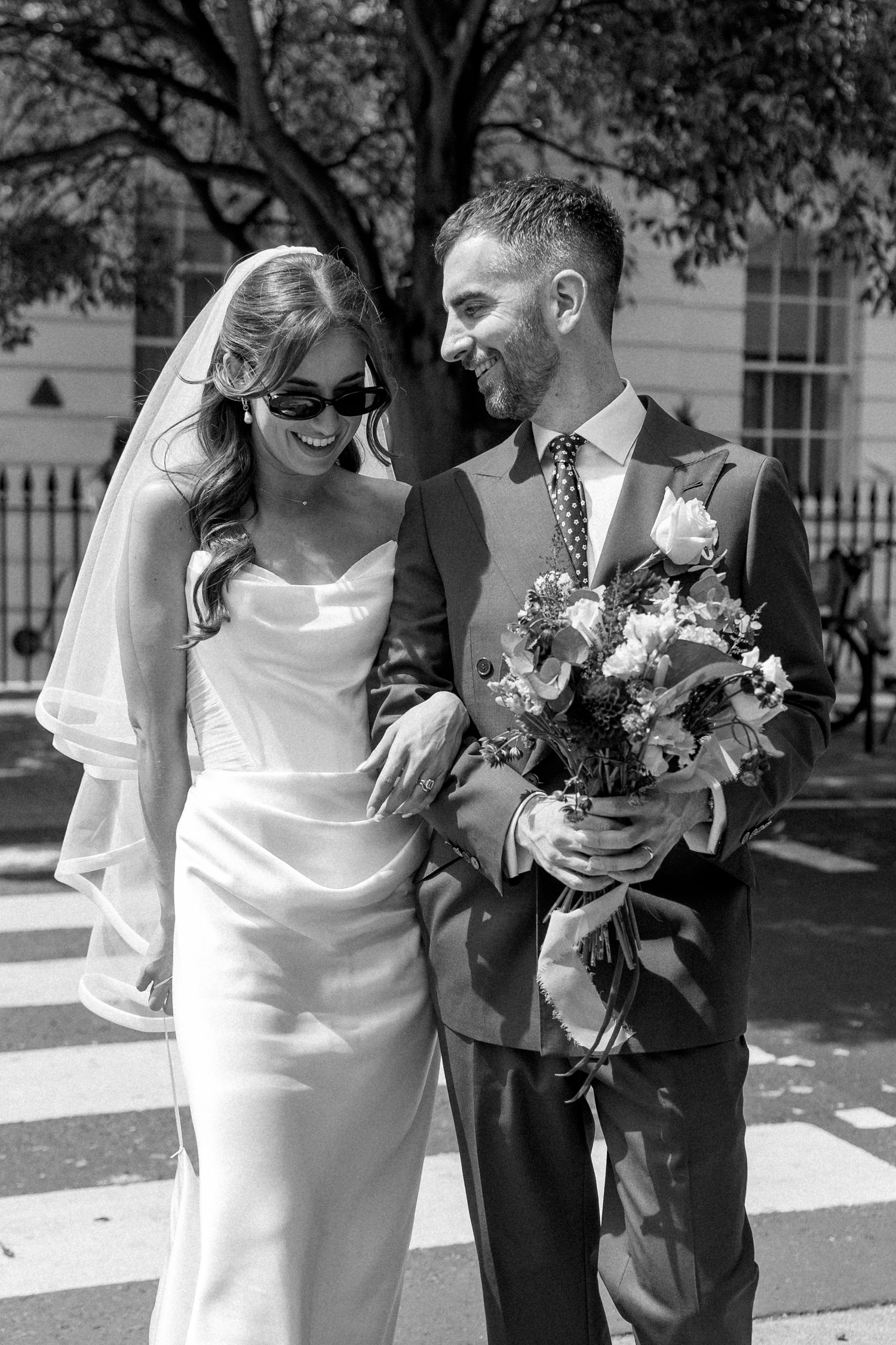 Smiling bride in white satin gown with veil and sunglasses walks with groom holding floral bouquet on a sunny street.