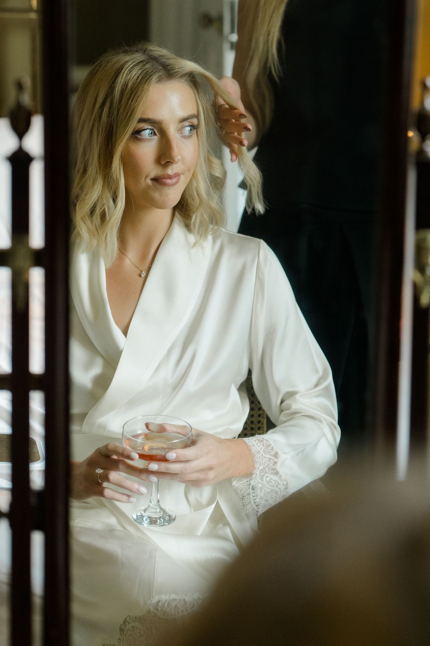 Blonde bride in white satin robe holding a cocktail glass while getting hair styled before wedding ceremony.