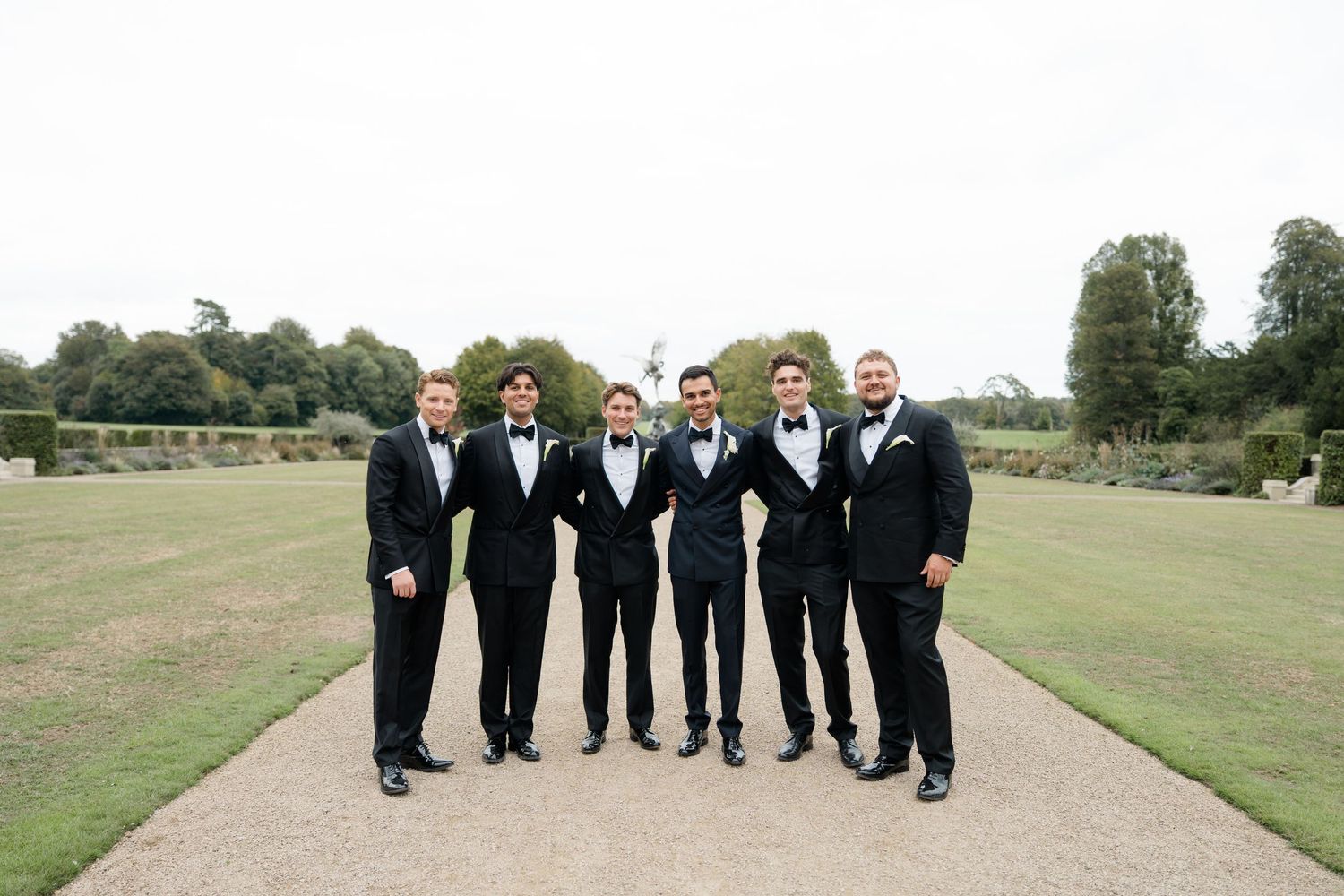 Six groomsmen pose together in matching black tuxedos on a garden pathway at a wedding.