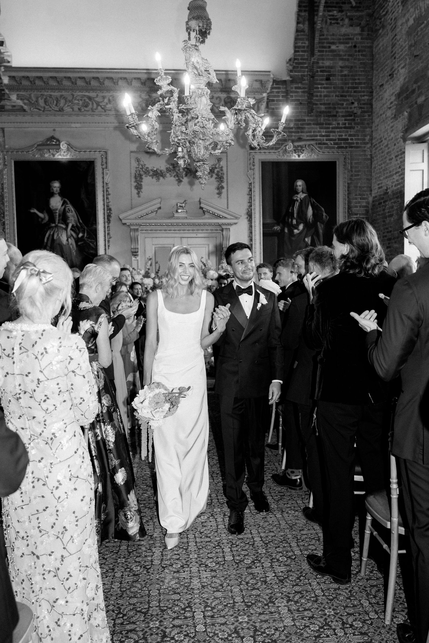 Black and white photo of bride and groom walking down aisle under chandelier with cheering guests.