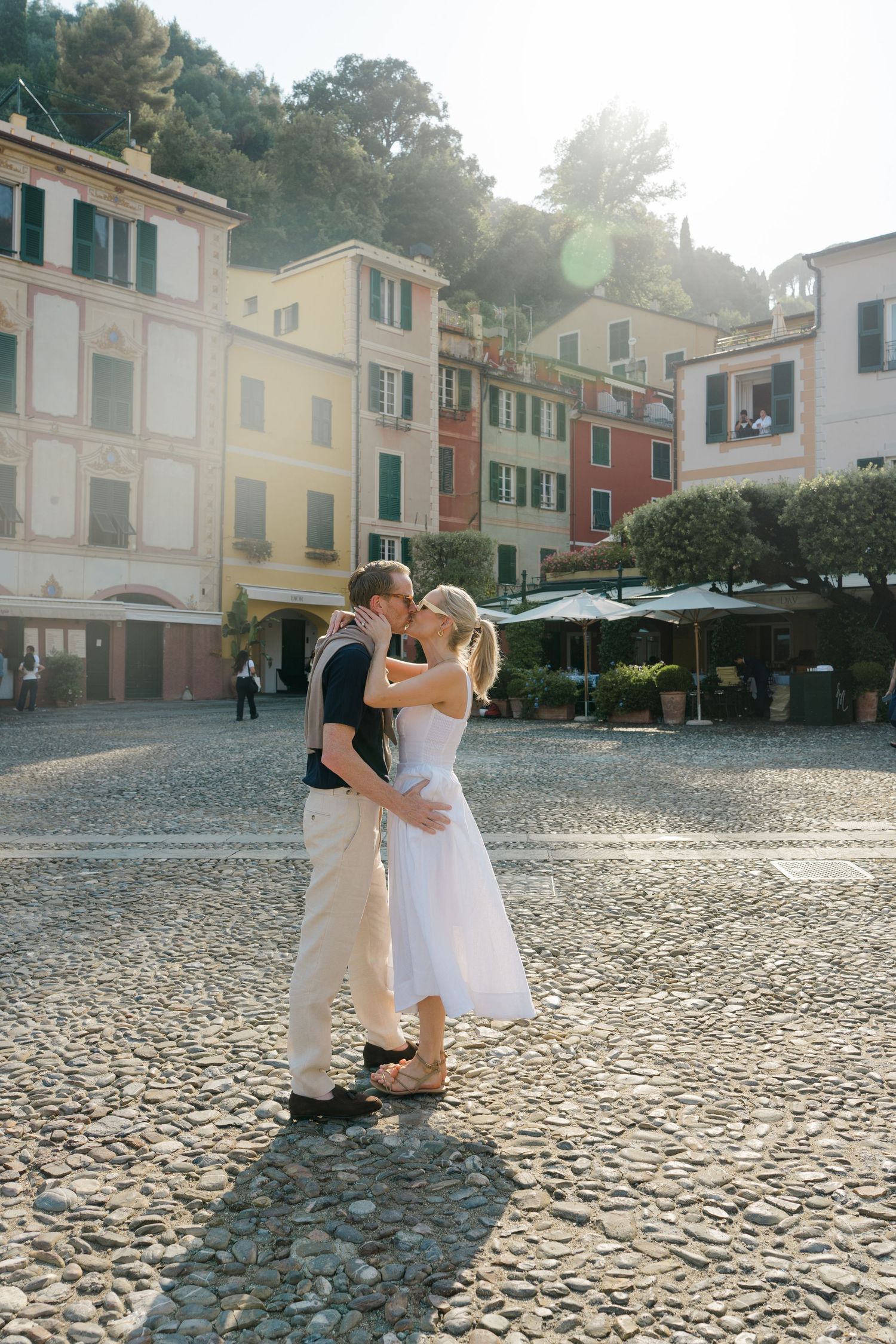 Couple sharing a romantic kiss in a sunlit cobblestone piazza in Portofino, Italy.
