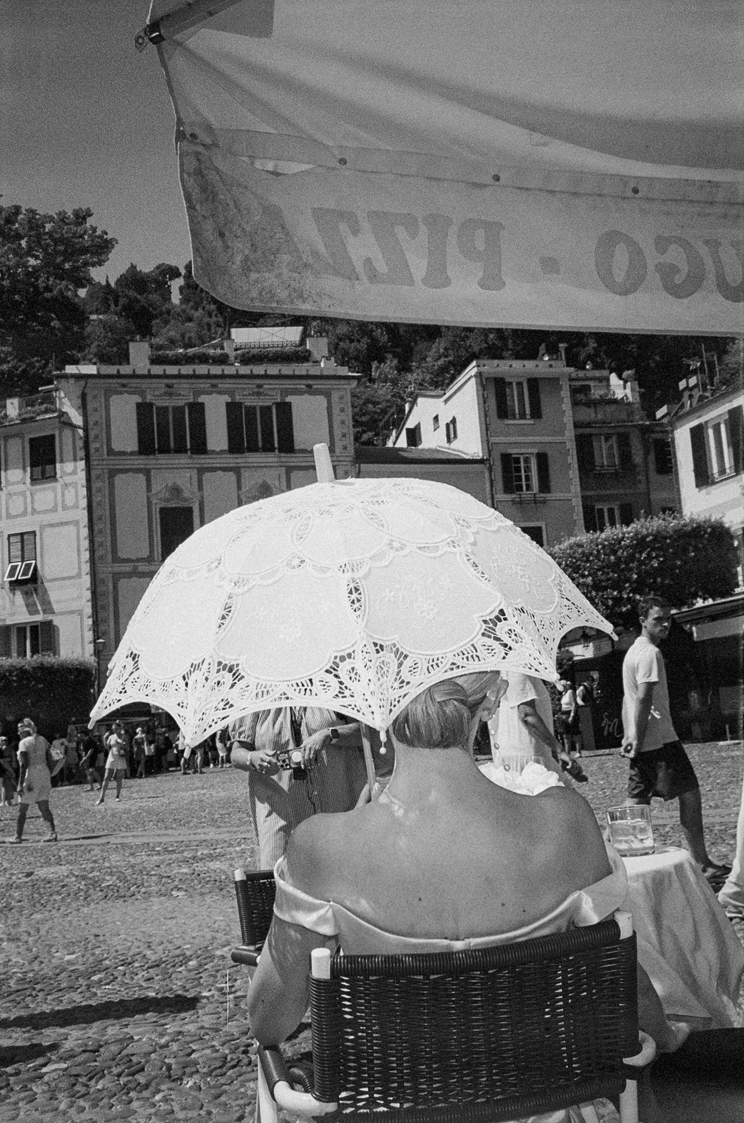 Black and white photo of person seated under ornate lace parasol in Italian coastal town square.