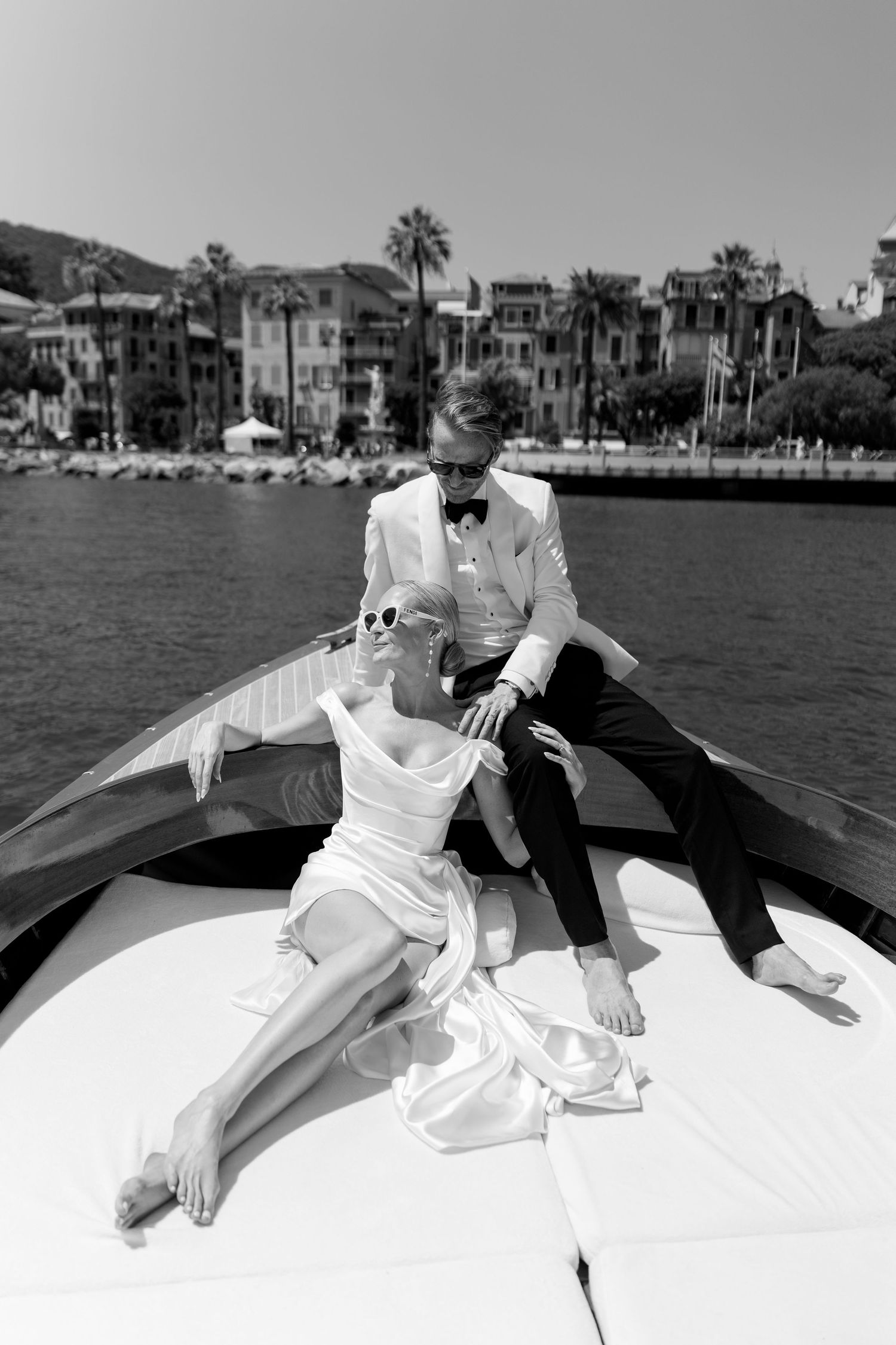 Bride and groom relaxing on bow of wooden boat with Italian coastal town in background.