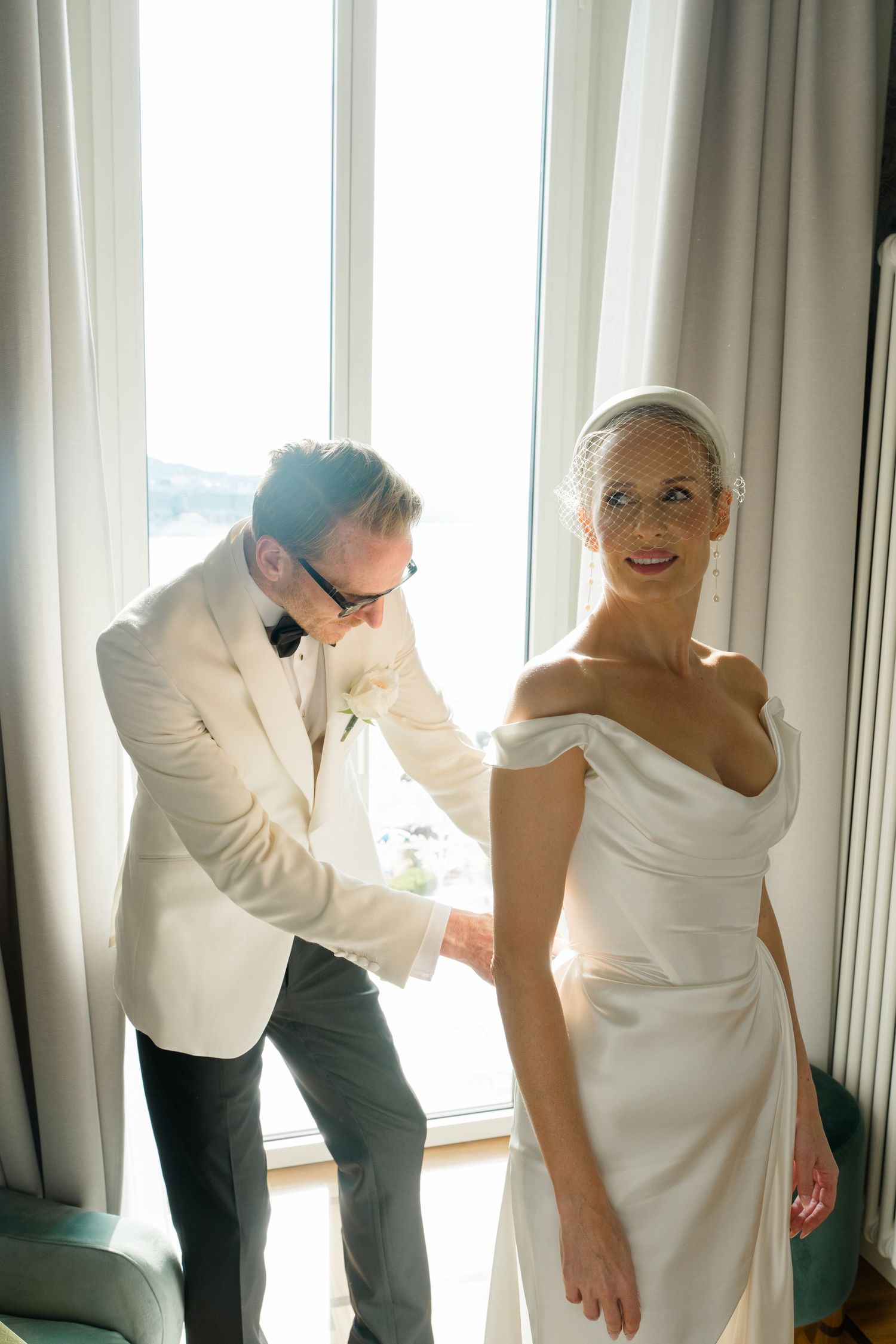 Groom in white tuxedo jacket helping bride in off-shoulder wedding dress near bright window.
