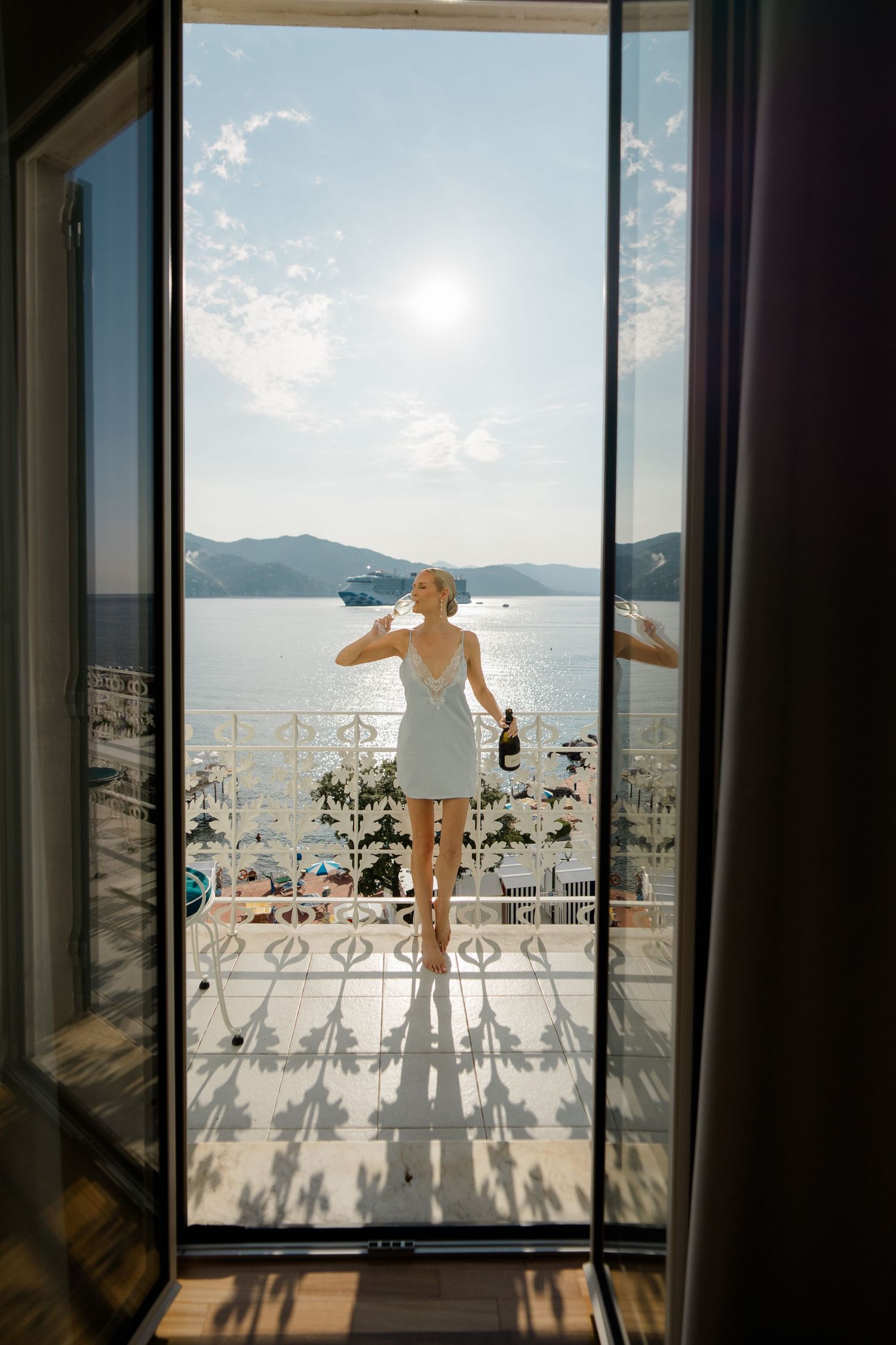 Woman in white dress holds champagne bottle on sun-drenched hotel balcony with stunning sea view.
