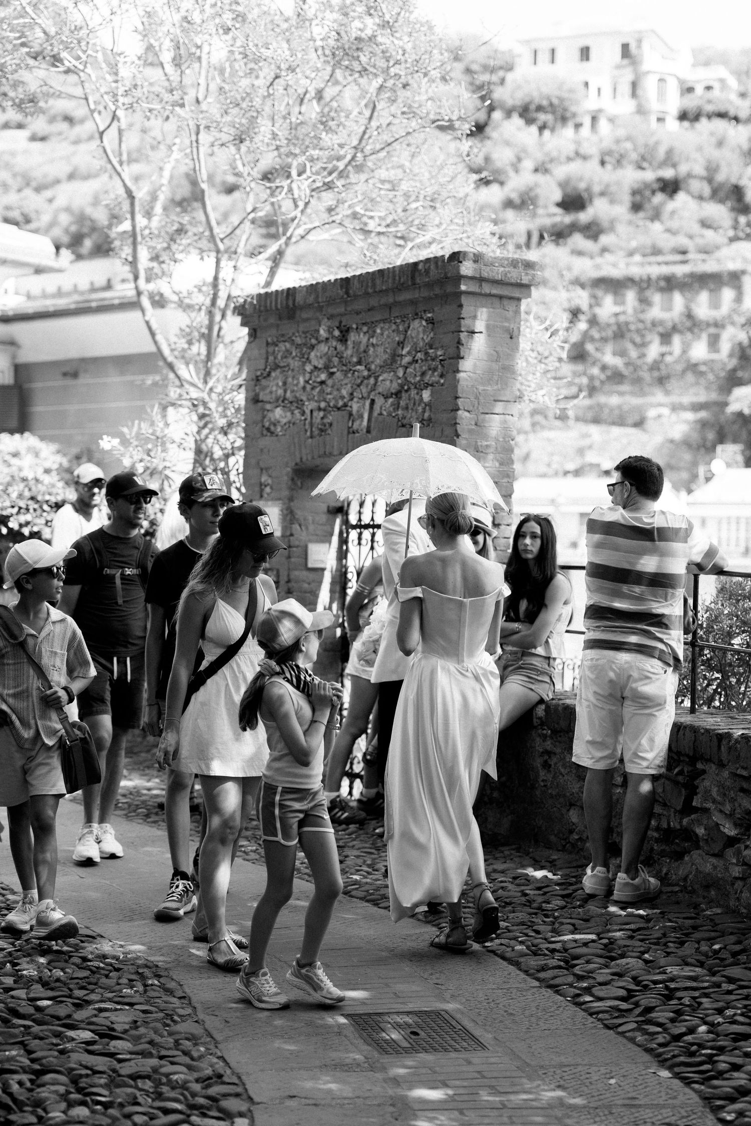 Bride in white gown with parasol walks among tourists on cobblestone path in black and white.