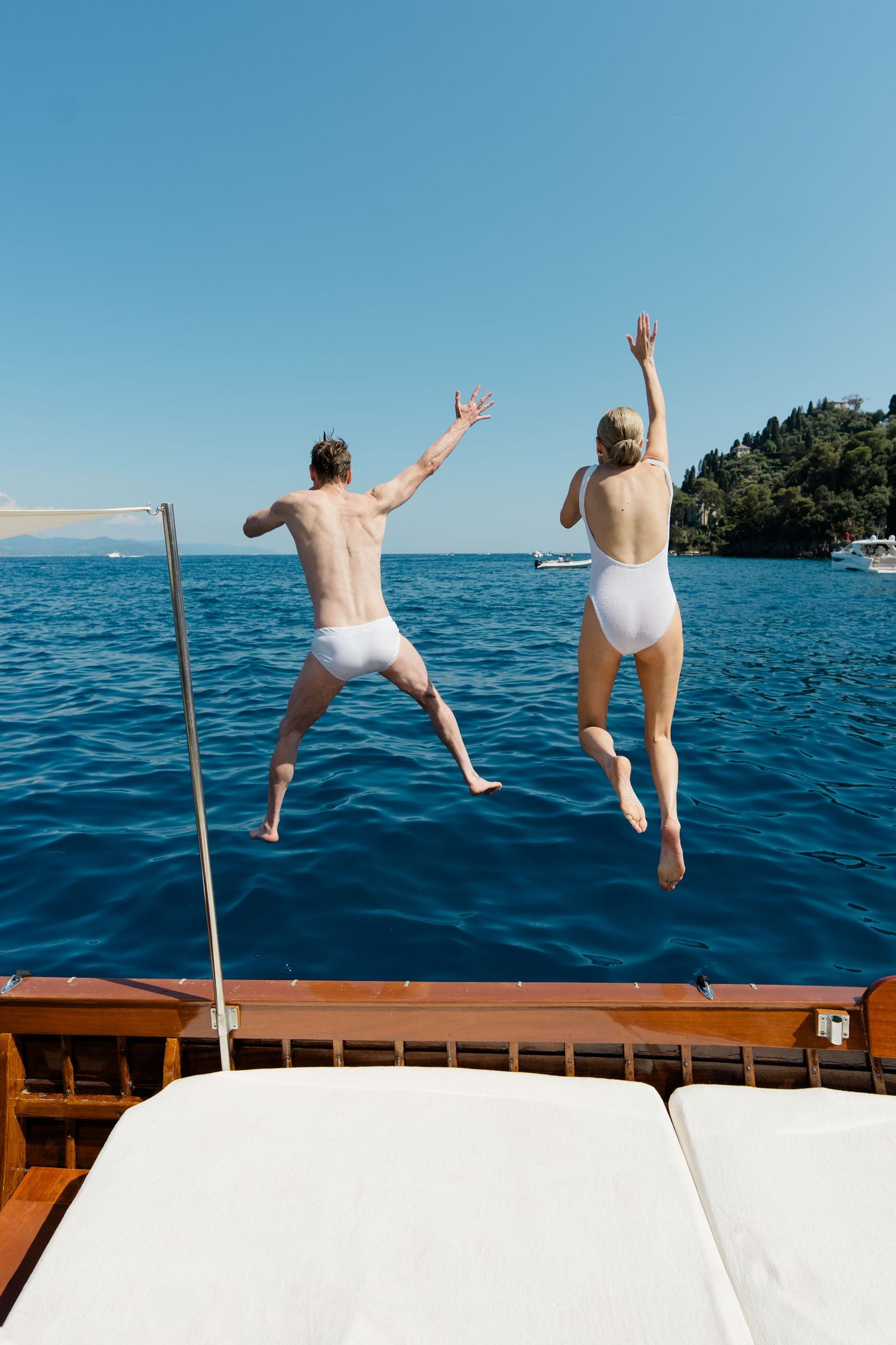 Bride and groom jumping off a wooden yacht into crystal blue Mediterranean waters in white swimwear.