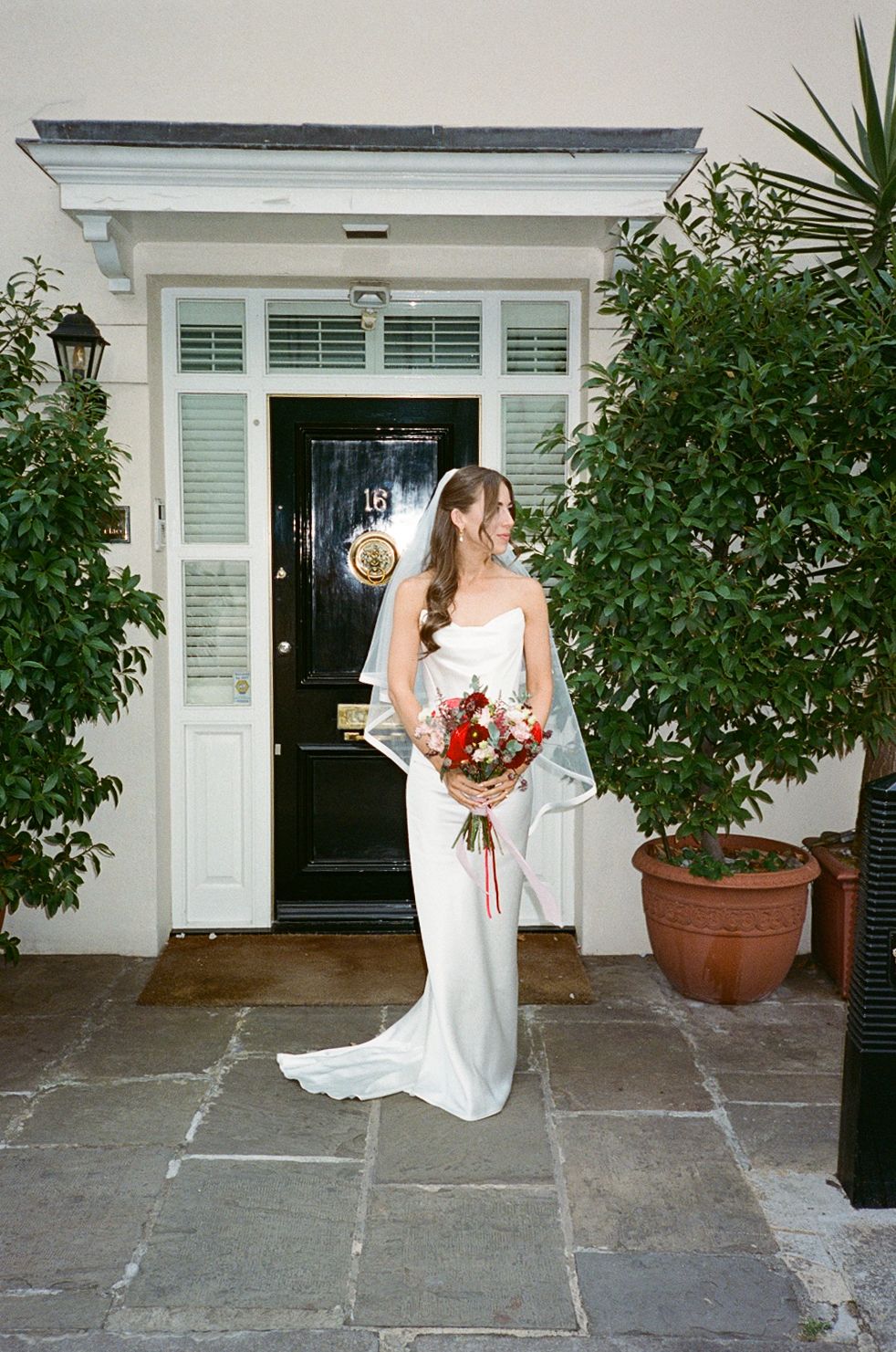 Elegant bride in white satin gown with veil holds red bouquet outside classic white Georgian townhouse doorway.