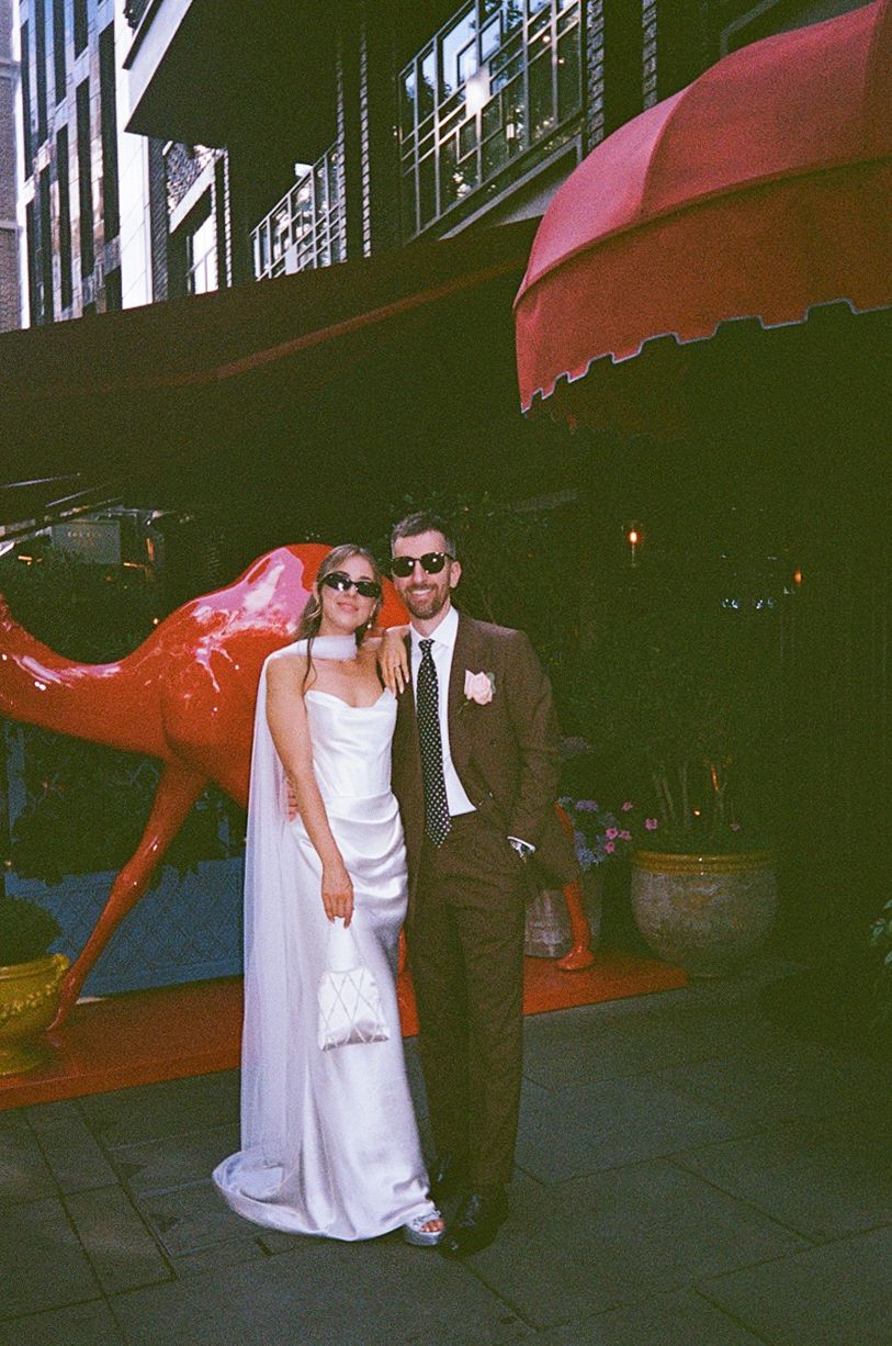 Bride and groom in sunglasses pose beside red sculpture outside upscale restaurant venue.
