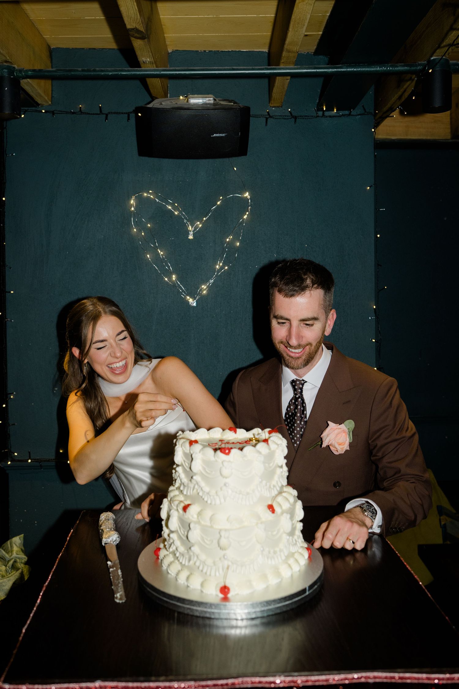 Bride and groom smiling while cutting a white tiered wedding cake with fairy light heart backdrop.