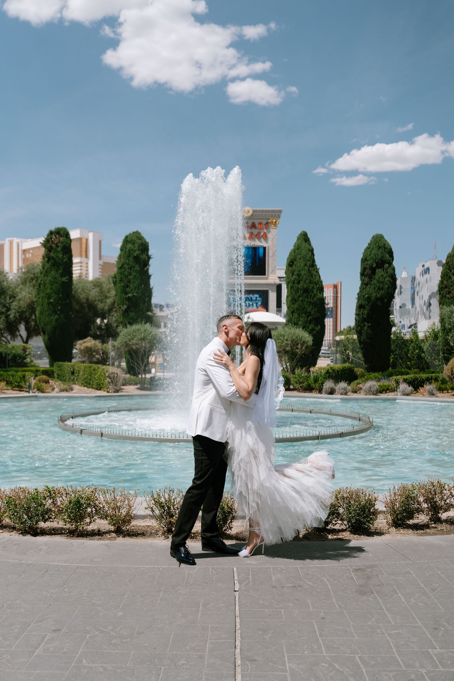 A romantic wedding photo in front of a large fountain with cypress trees and blue sky.