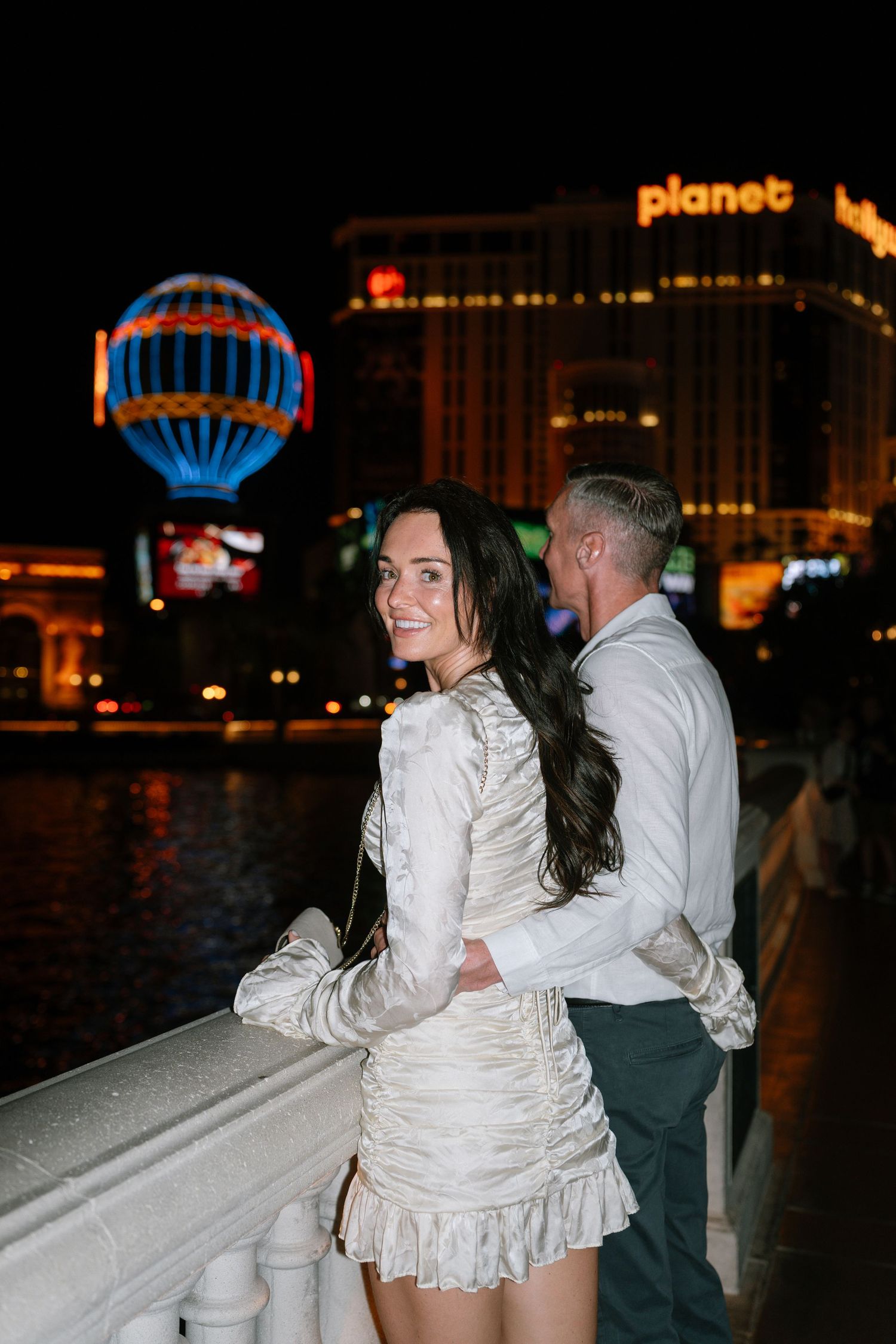 A couple poses against a balustrade with the iconic Las Vegas strip lights in the background.