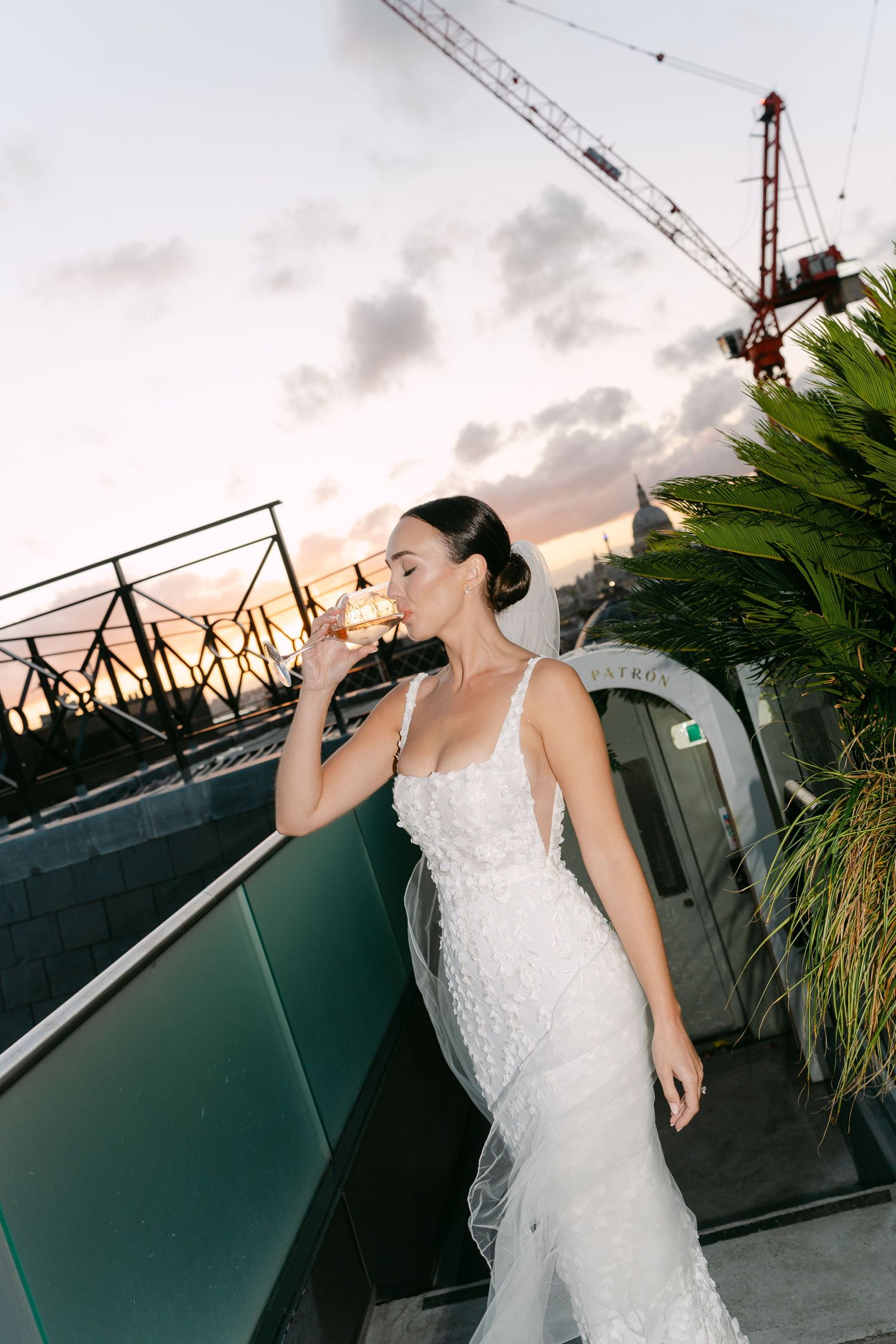 Elegant white lace wedding dress against sunset sky with construction cranes visible.