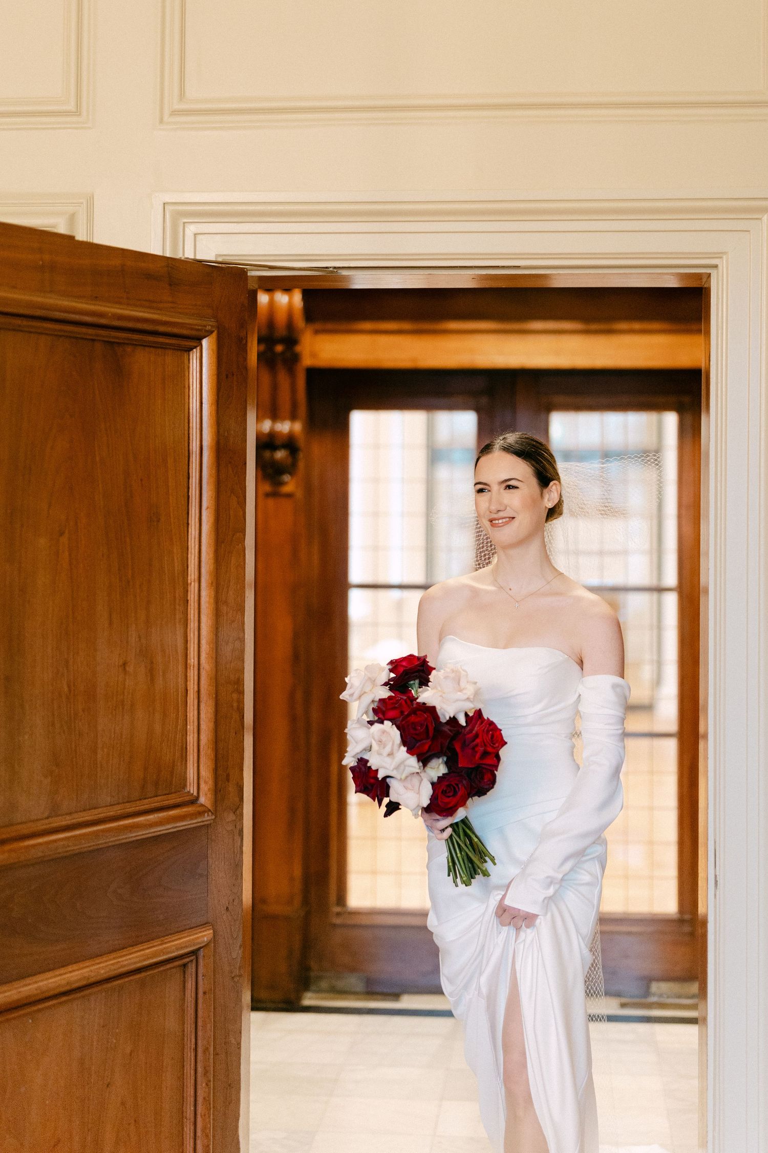 A bride in a white gown holds red and white flowers while standing in an elegant wood-paneled hallway.