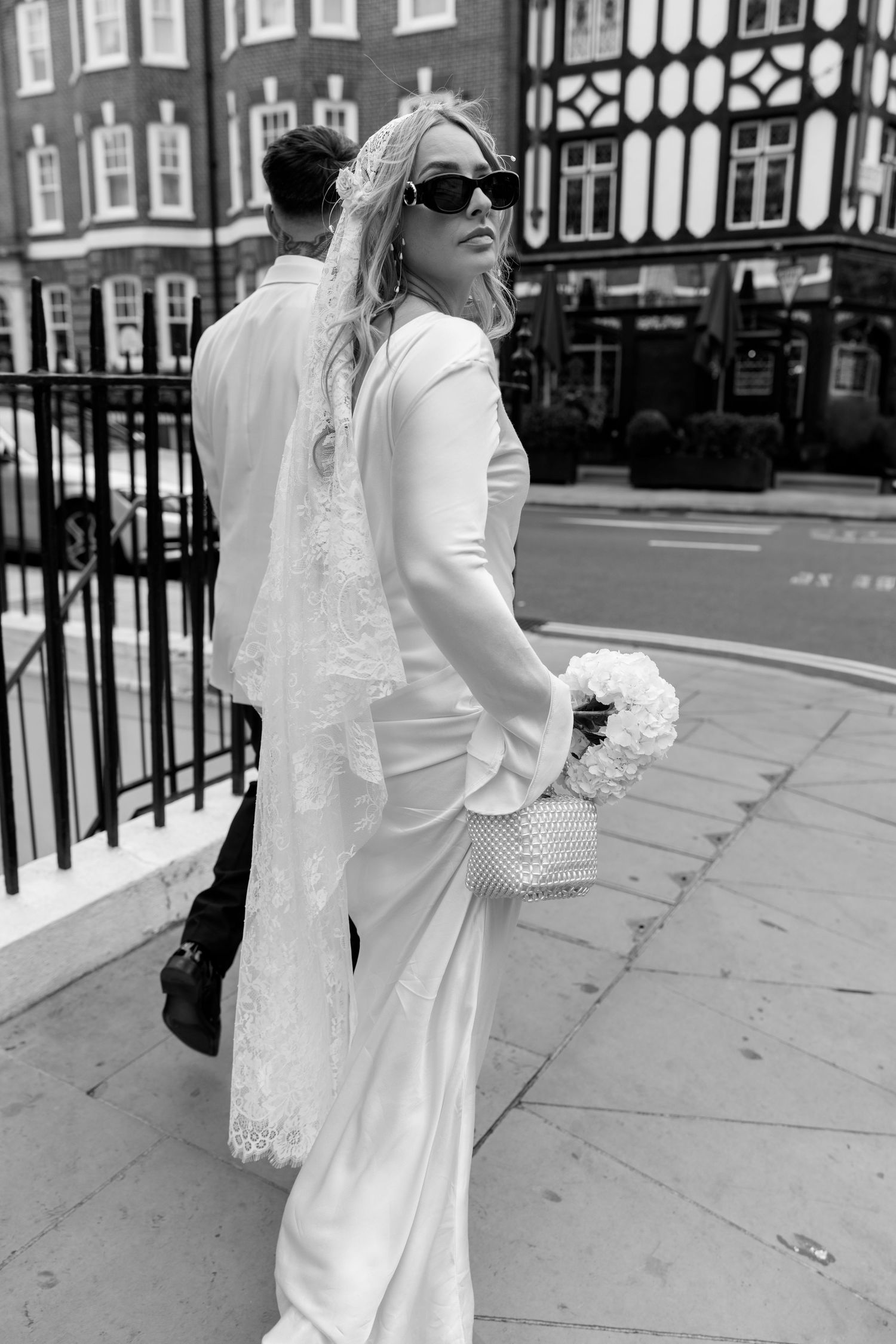 Black and white photo of a bride in a white dress walking along an iron-fenced sidewalk.