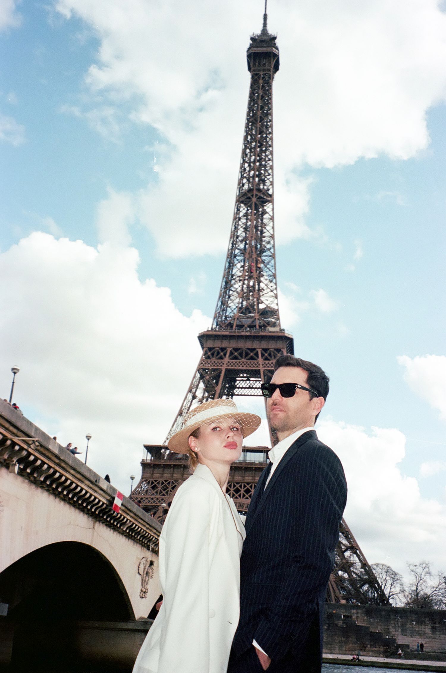A stylish couple poses in front of the iconic Eiffel Tower in Paris on a bright, cloudy day.