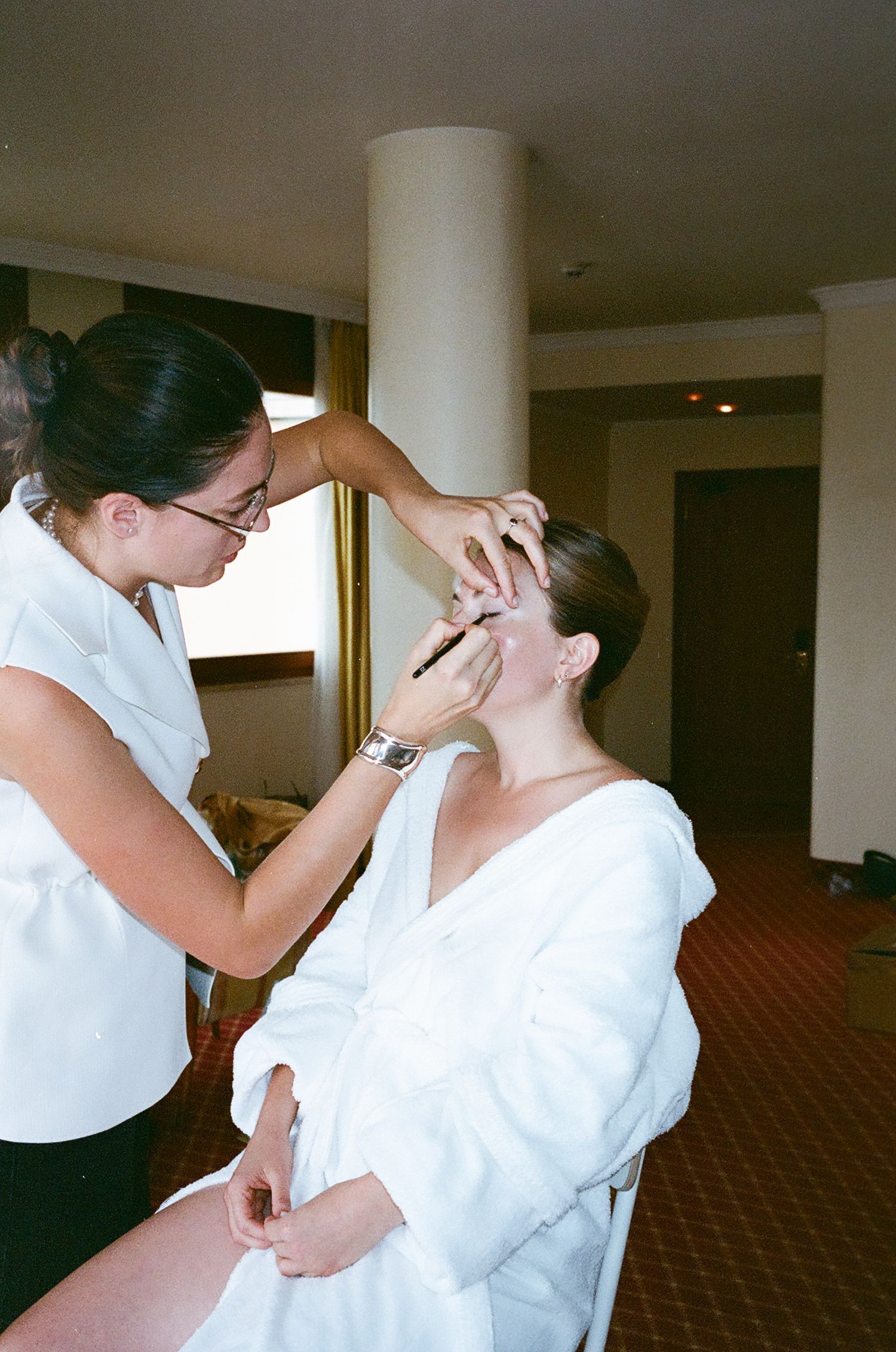 A makeup artist applies cosmetics to a client wearing a white robe in a hotel room setting.