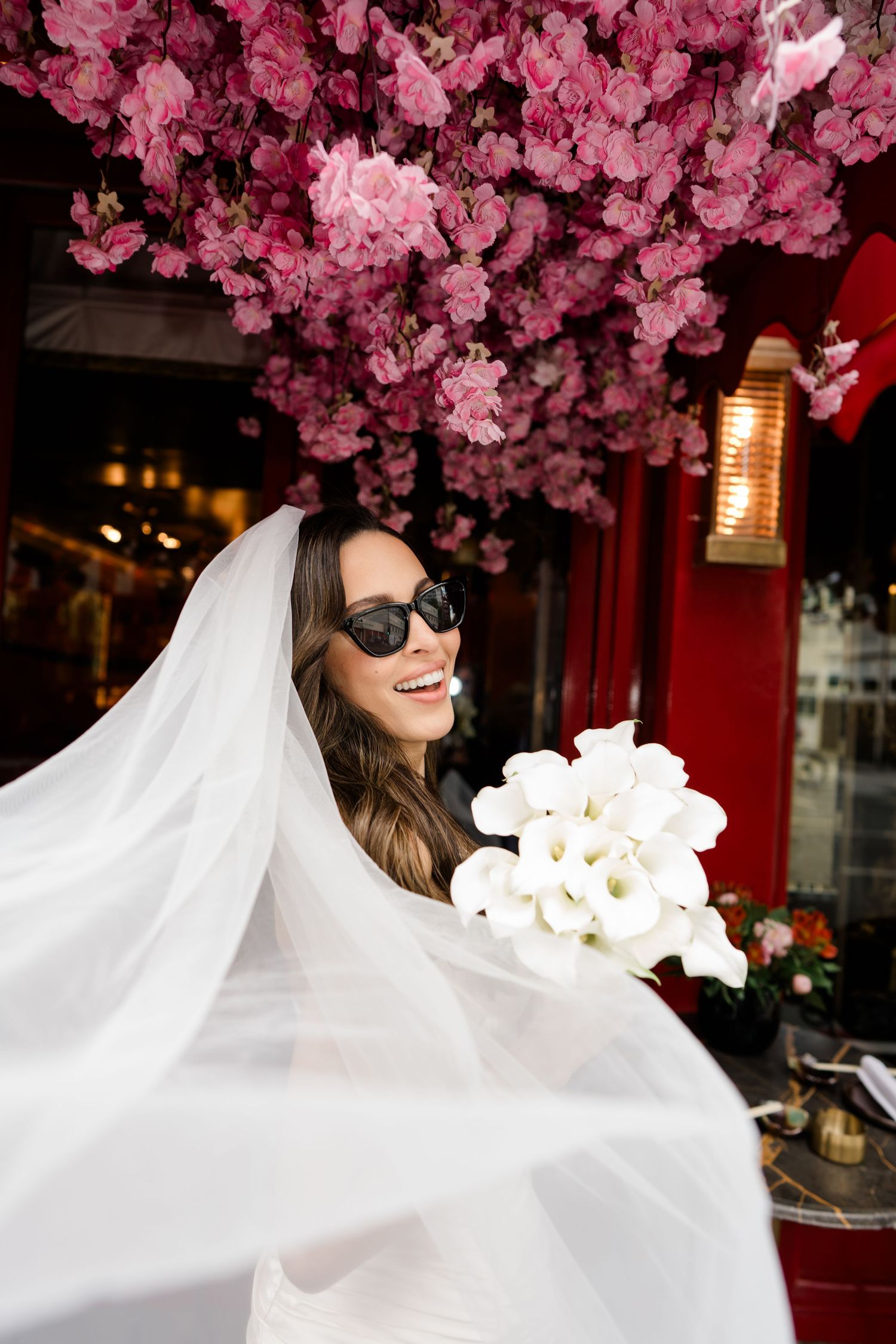 A bride in sunglasses stands under pink cherry blossoms with a flowing white veil against a red building.