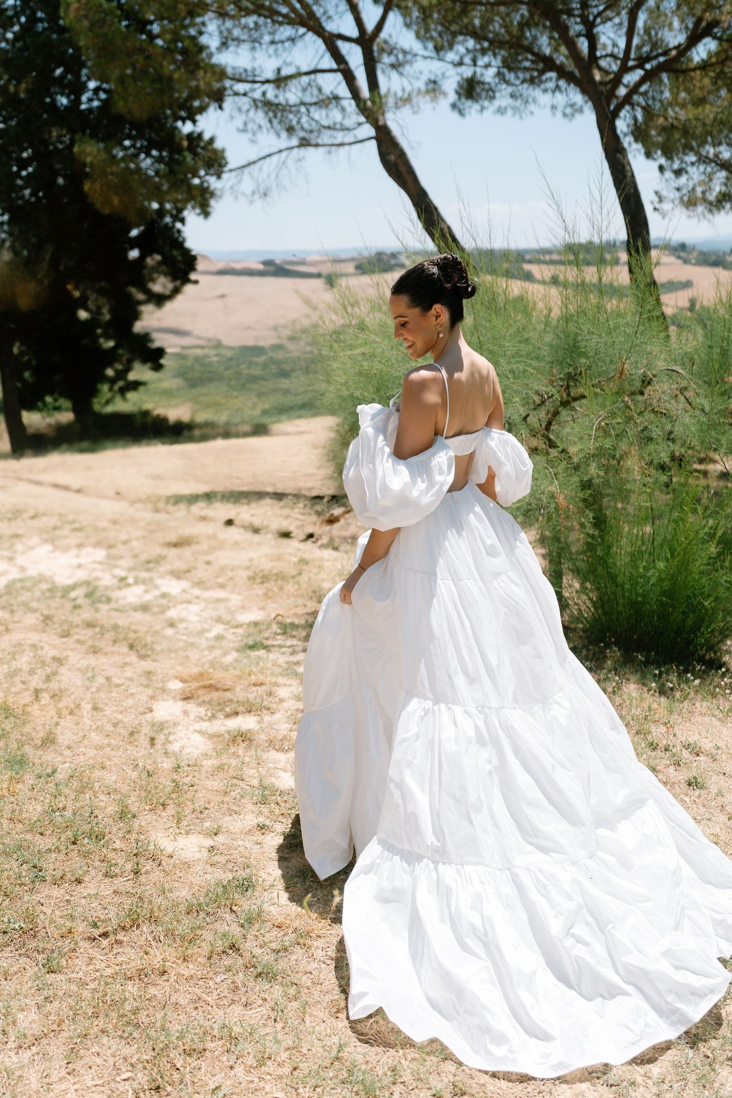 A flowing white wedding dress billows in the wind as someone twirls on a grassy hilltop on a sunny day.