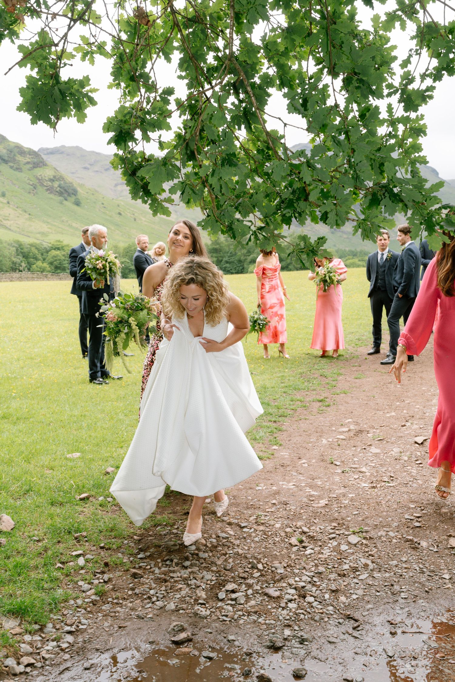 Wedding party walking through grassy field under tree branches in outdoor setting.