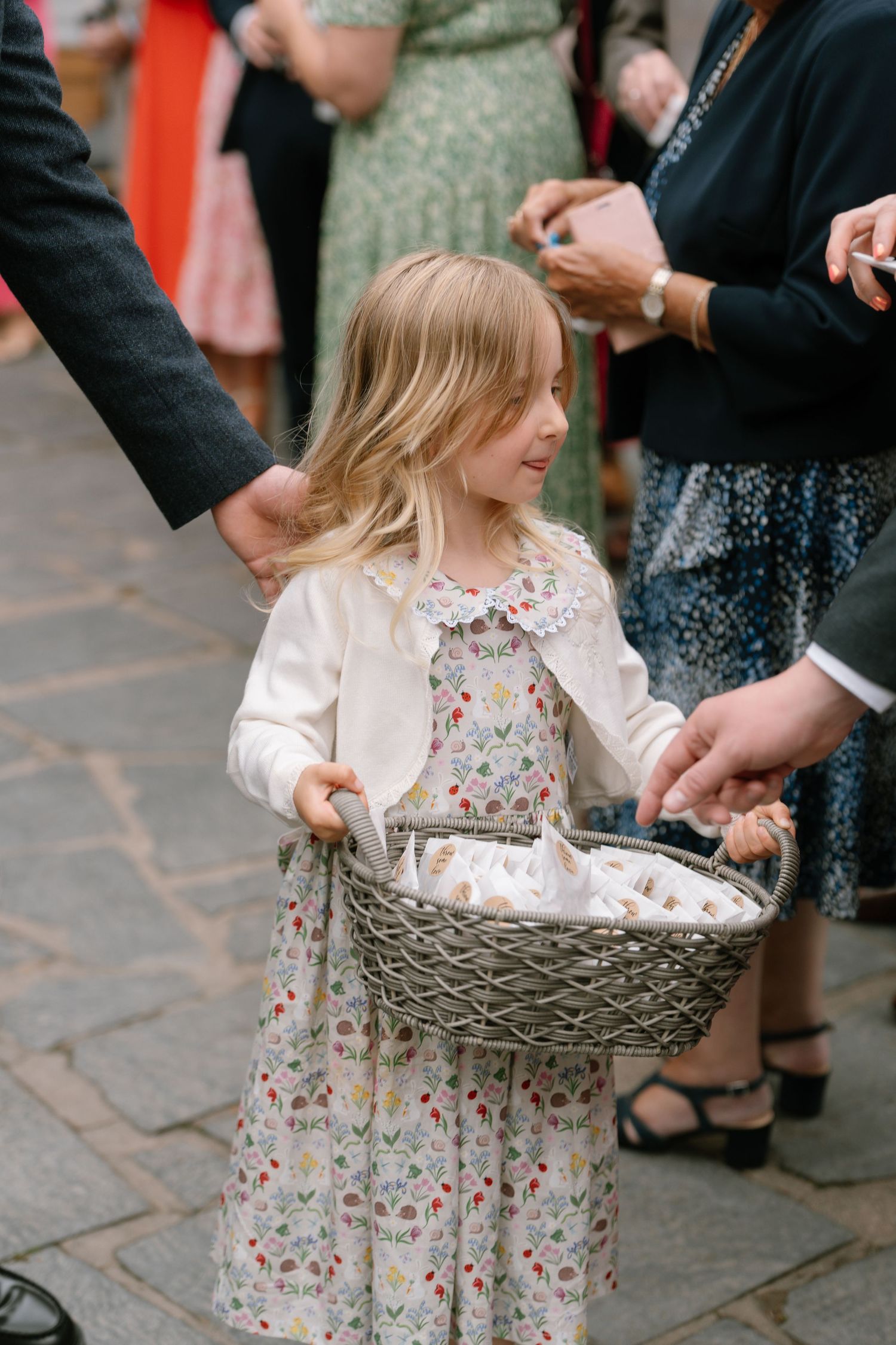 Young flower girl in floral dress playing with basket at outdoor wedding.