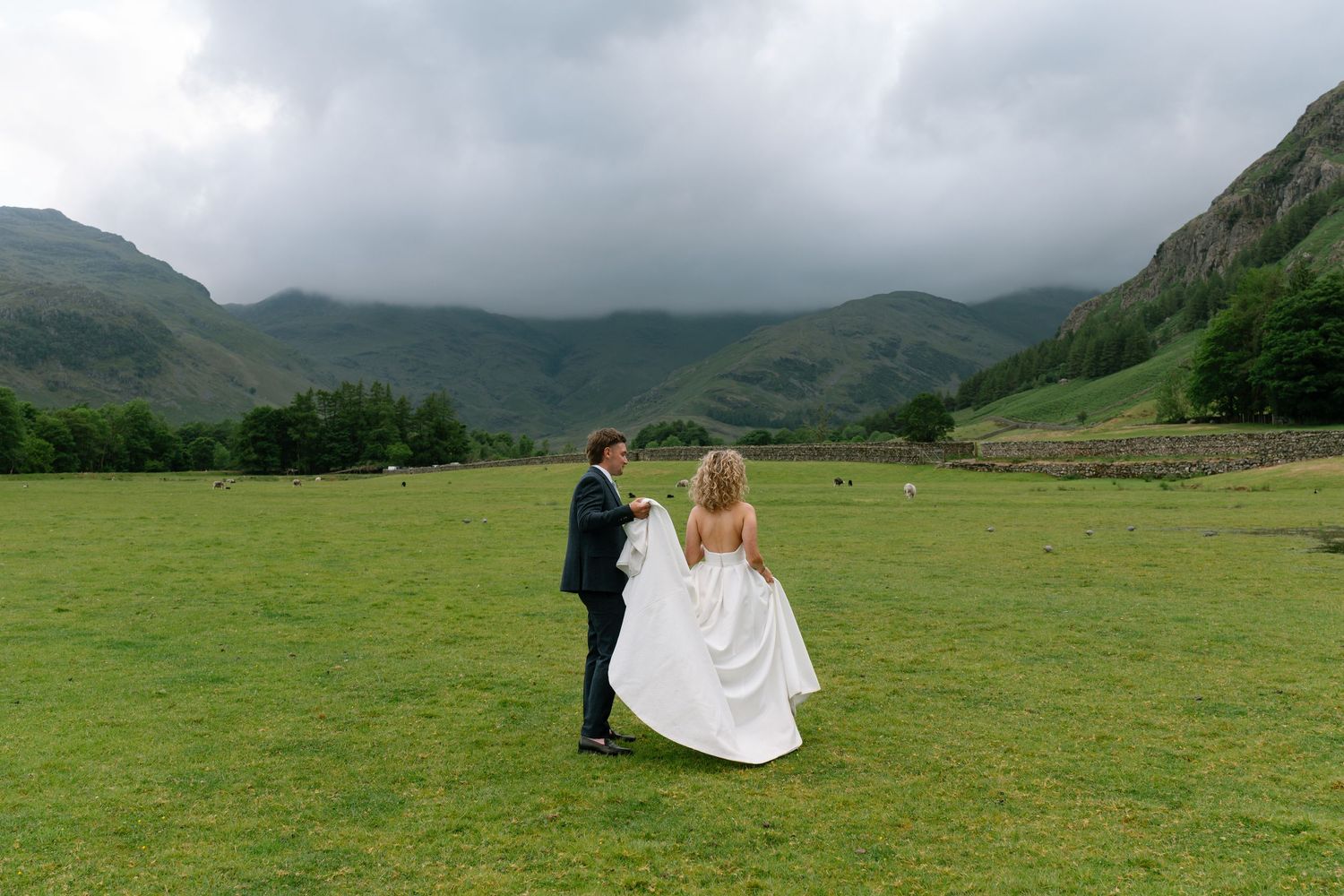 Couple walking through a green field with dramatic mountain scenery and cloudy skies.