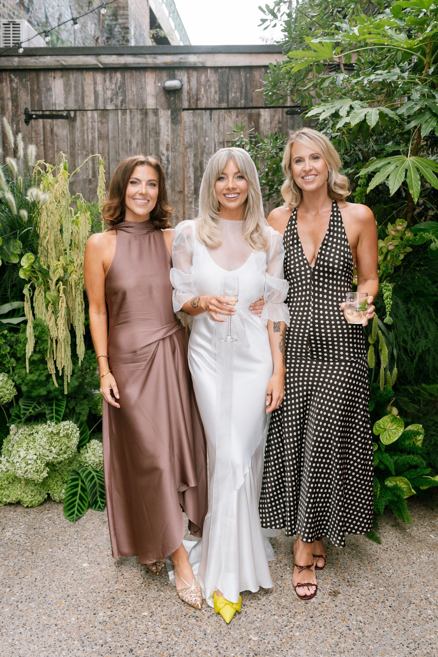 Three wedding attendees pose together in elegant dresses at outdoor garden reception.