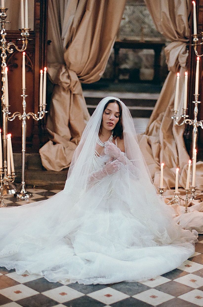 Bride in flowing white wedding gown with long veil poses dramatically on marble floor with candlesticks.