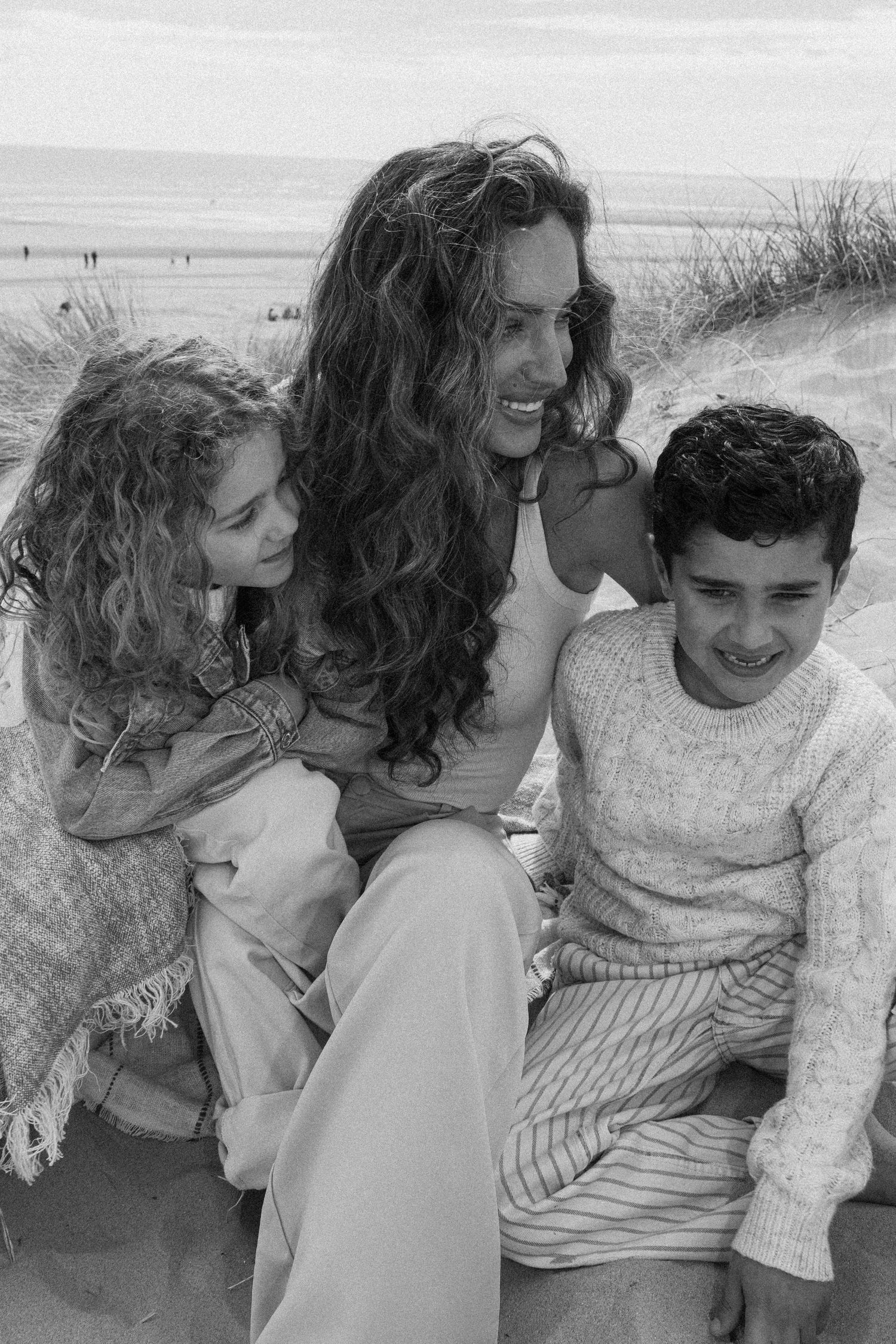 Black and white photo of a woman with curly hair sitting on beach sand with two children, smiling warmly.