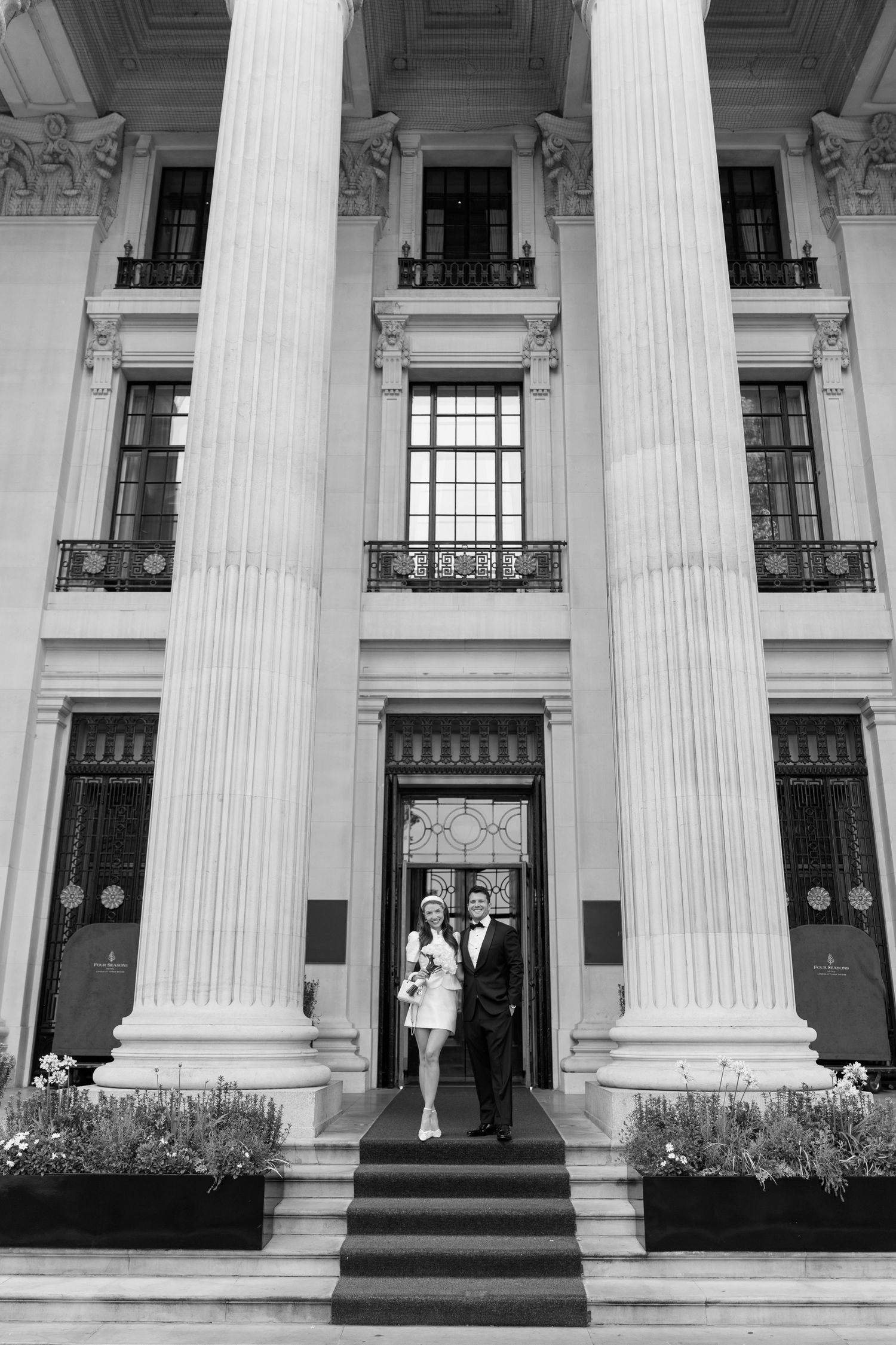 Black and white photo of a couple in wedding attire posing on steps of a grand neoclassical building with tall columns.