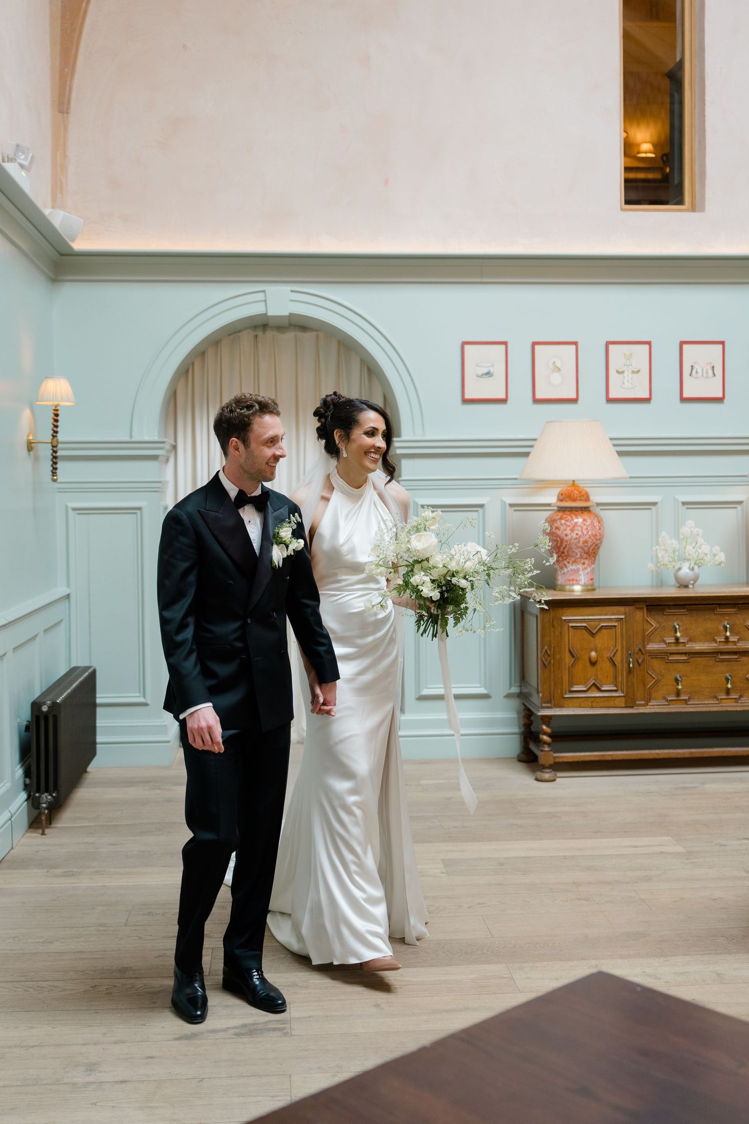 Bride in white halter gown and groom in black tuxedo walking through an elegant blue-paneled reception room.