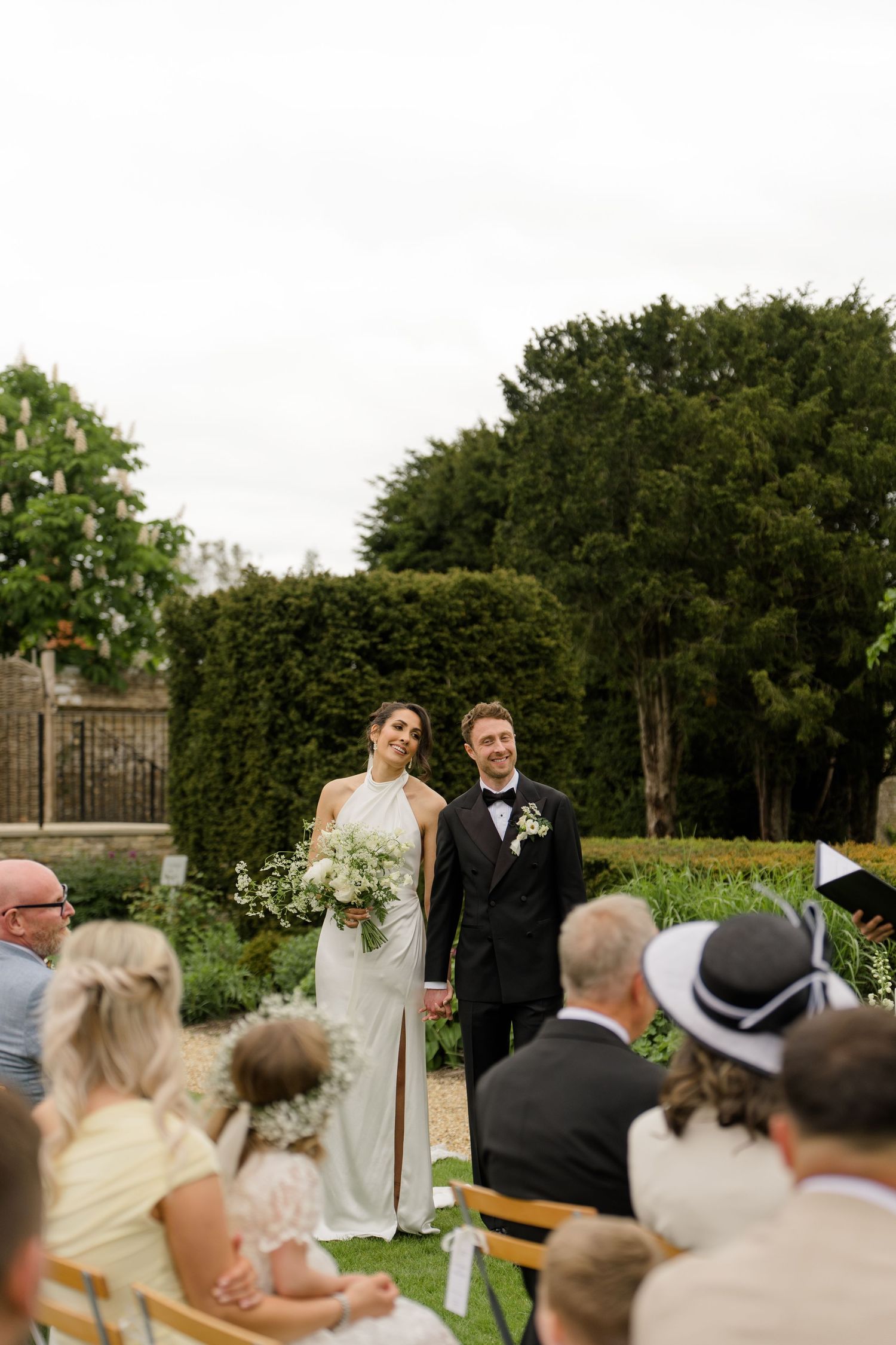 A smiling bride and groom stand together after their outdoor garden wedding ceremony surrounded by seated guests.