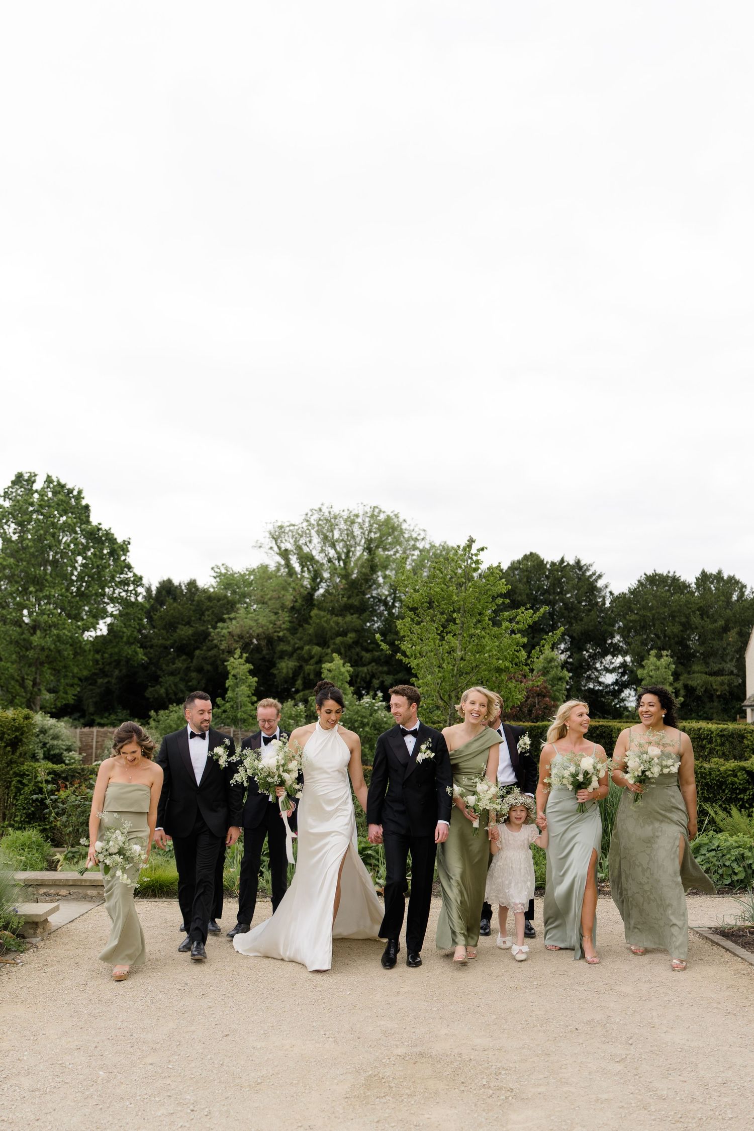 Wedding party walks together outdoors, bride in white gown, groomsmen in black tuxedos, bridesmaids in sage green dresses.