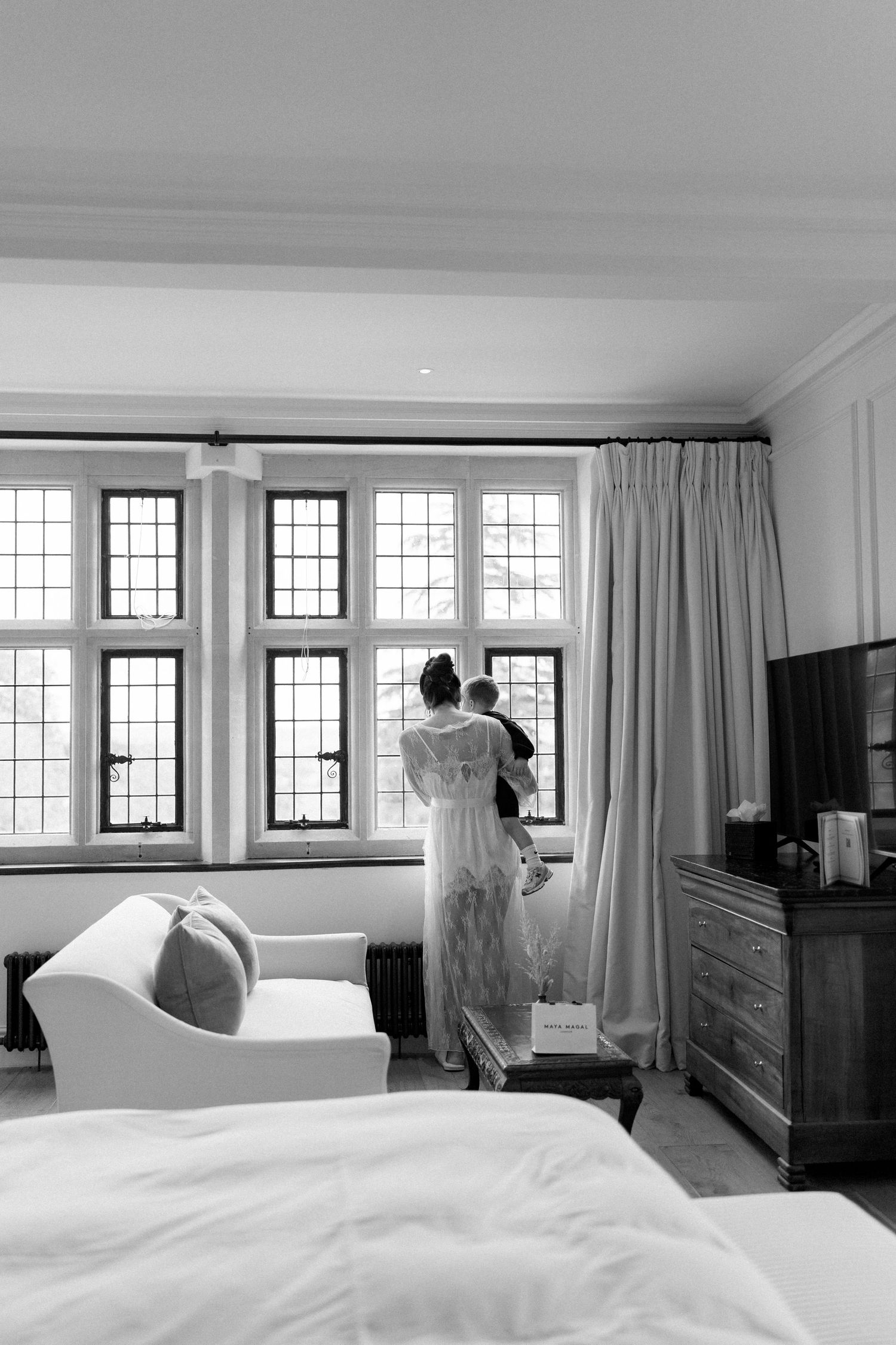 Black and white photo of bride in wedding dress holding baby while looking out large leaded glass windows.