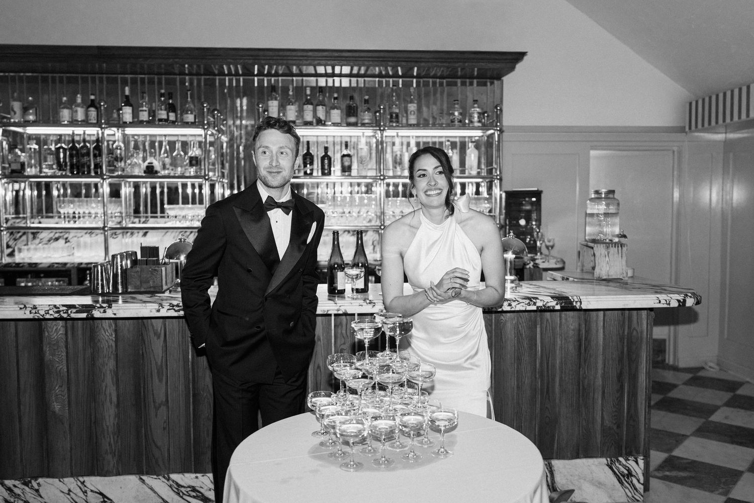 Black and white photo of bride and groom standing at champagne tower beside a well-stocked bar.