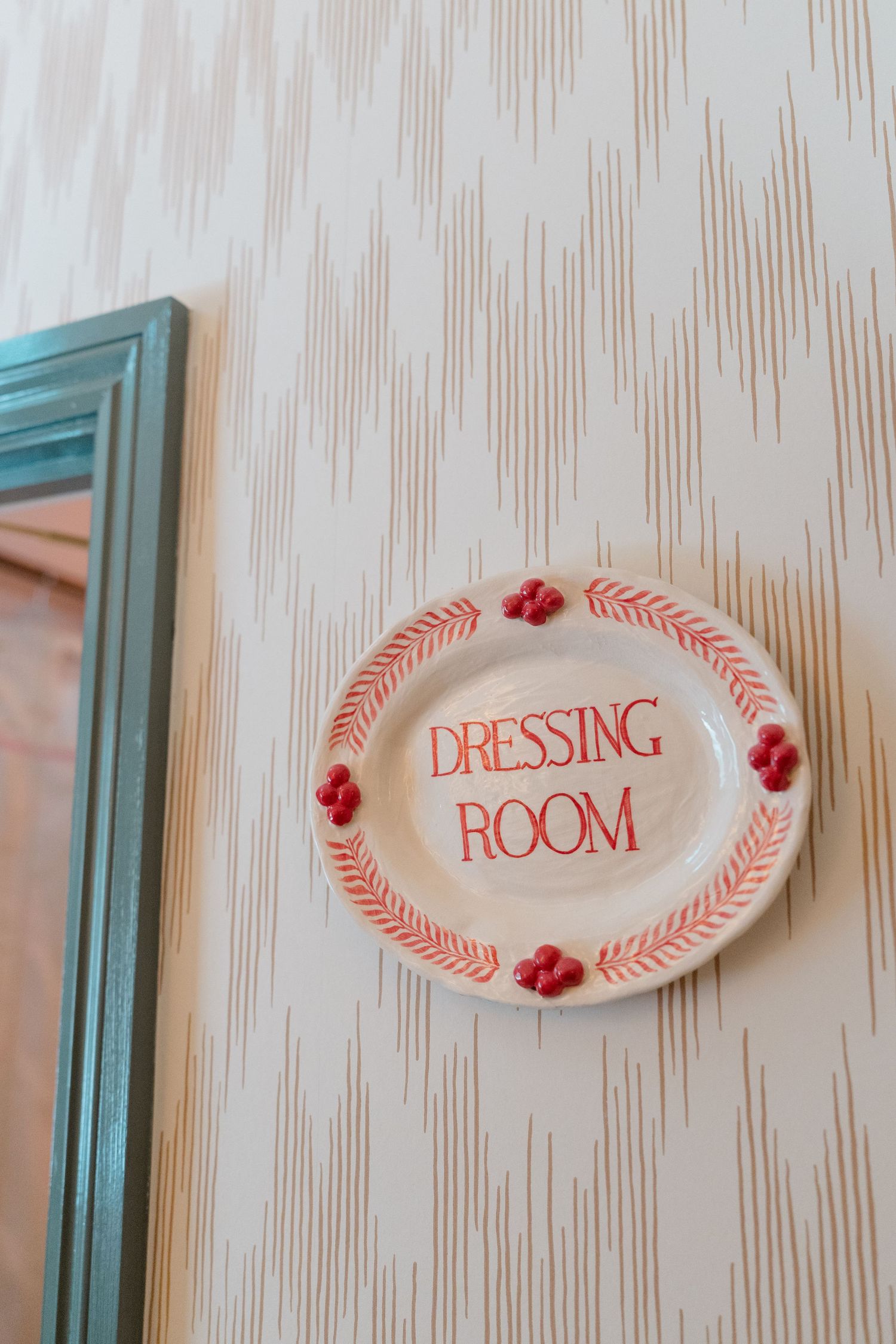 Decorative ceramic plate sign reading Dressing Room with red floral details mounted on striped wallpaper.