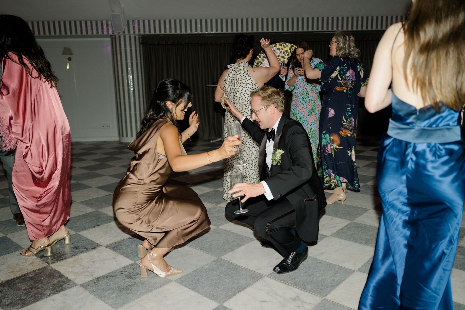 A bride in a white gown and a guest in bronze crouch down together on the dance floor at a wedding reception.