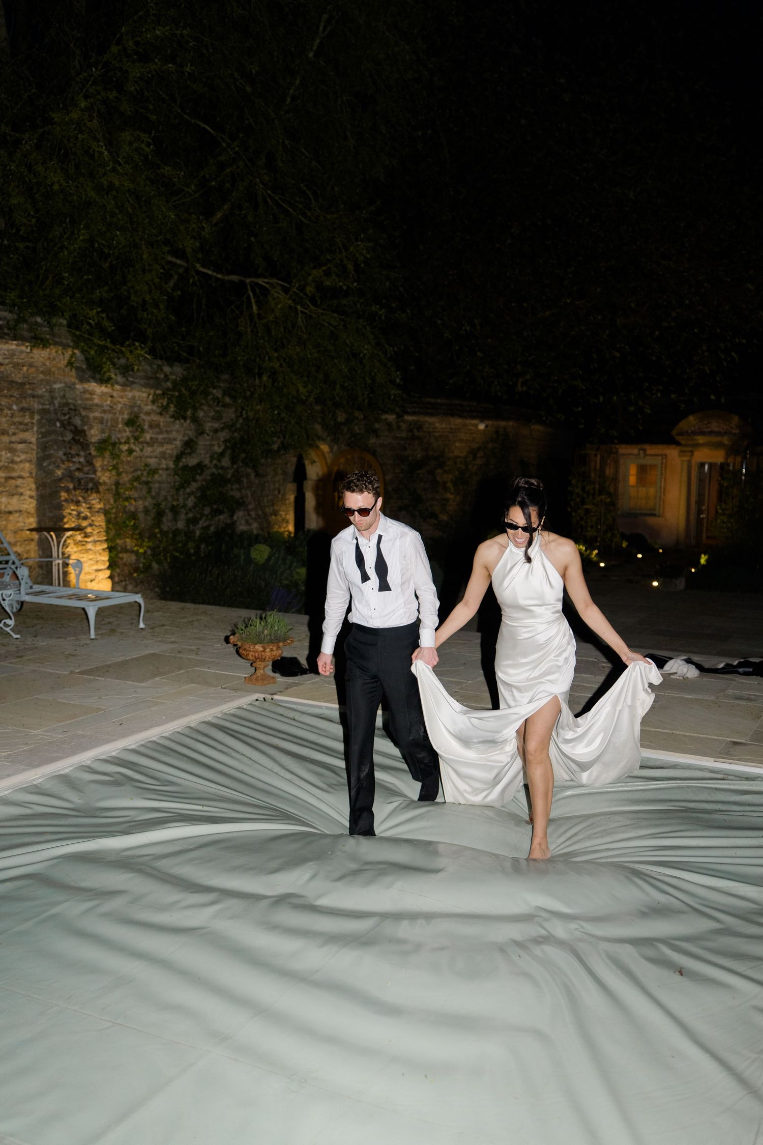 Bride and groom walk barefoot at night on outdoor dance floor, bride lifting white gown hem elegantly.