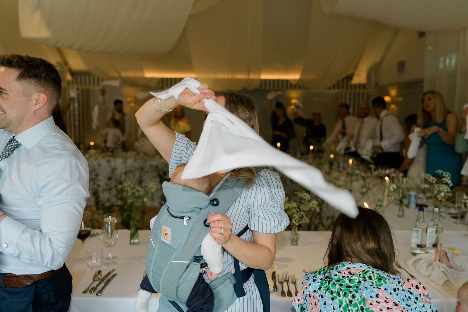 Guest wearing baby carrier playfully covers infant with a cloth napkin at wedding reception.