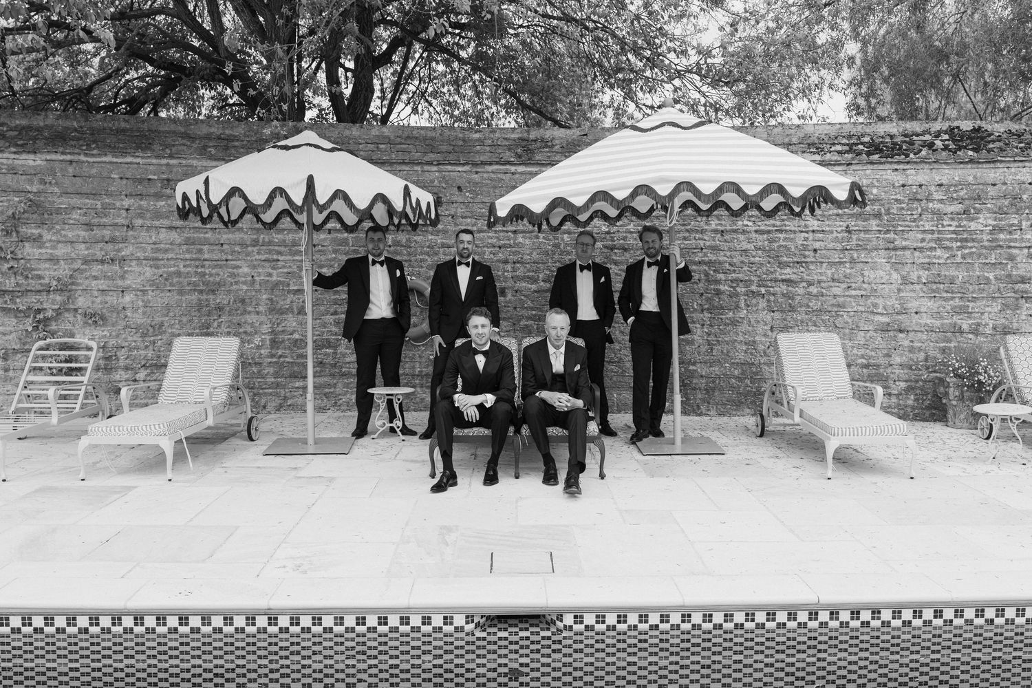 Groom and four groomsmen in black tuxedos posing by a pool under striped umbrellas.