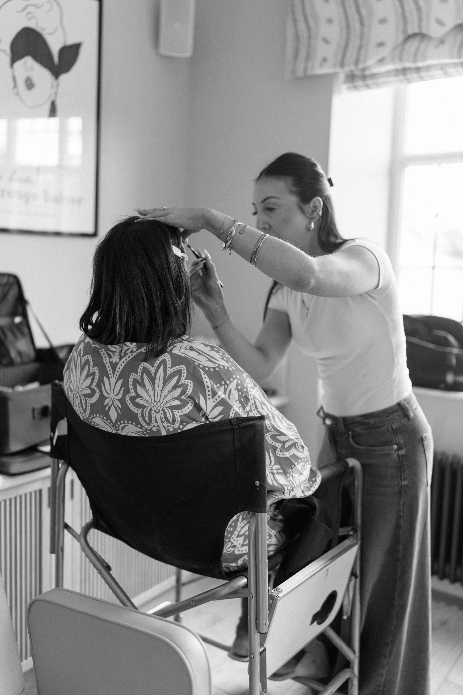 Black and white photo of a hairstylist working on a seated client's hair in a bright room.