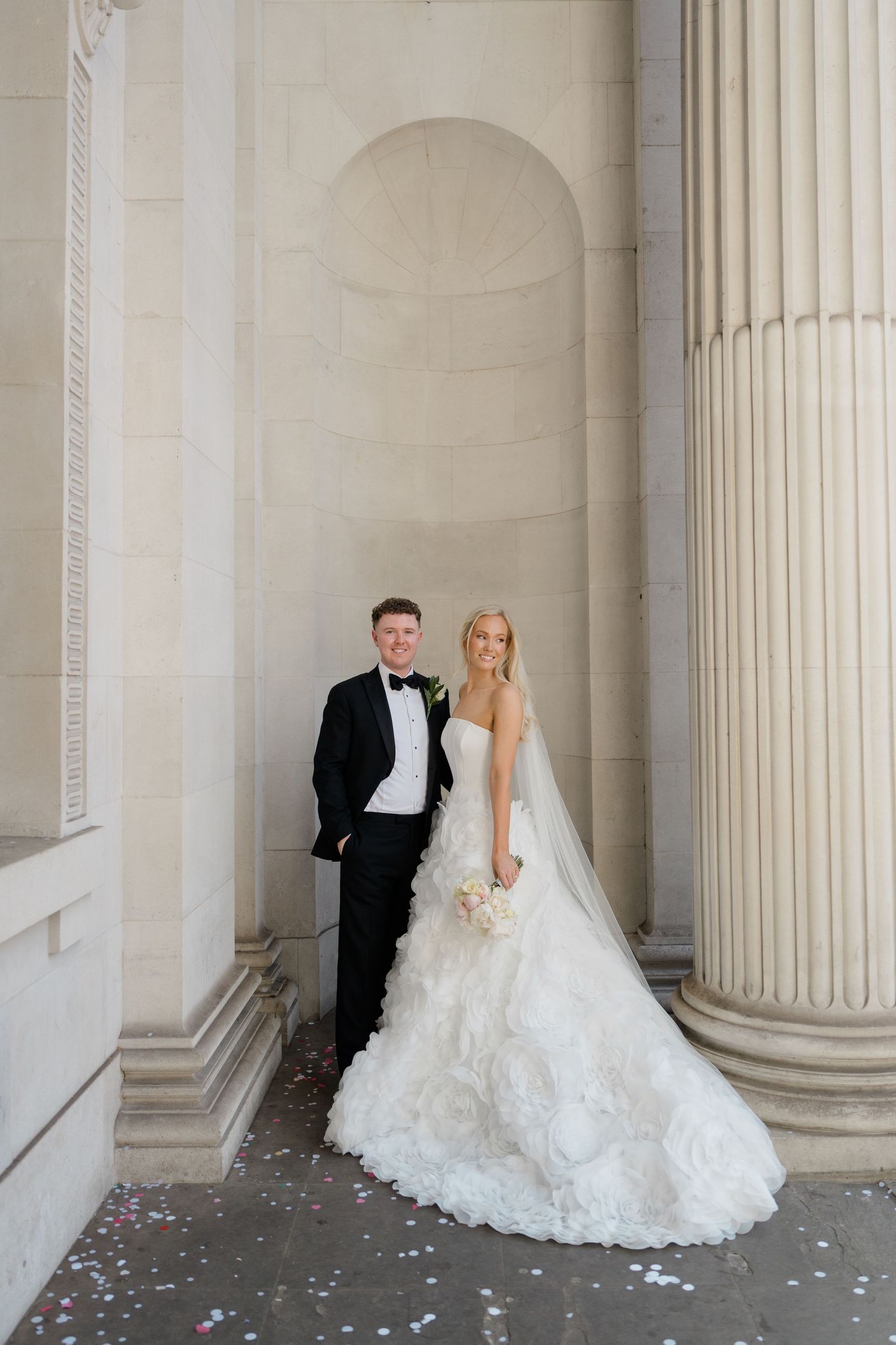 Bride in strapless ruffled gown and groom in black tuxedo pose among grand stone columns with confetti.