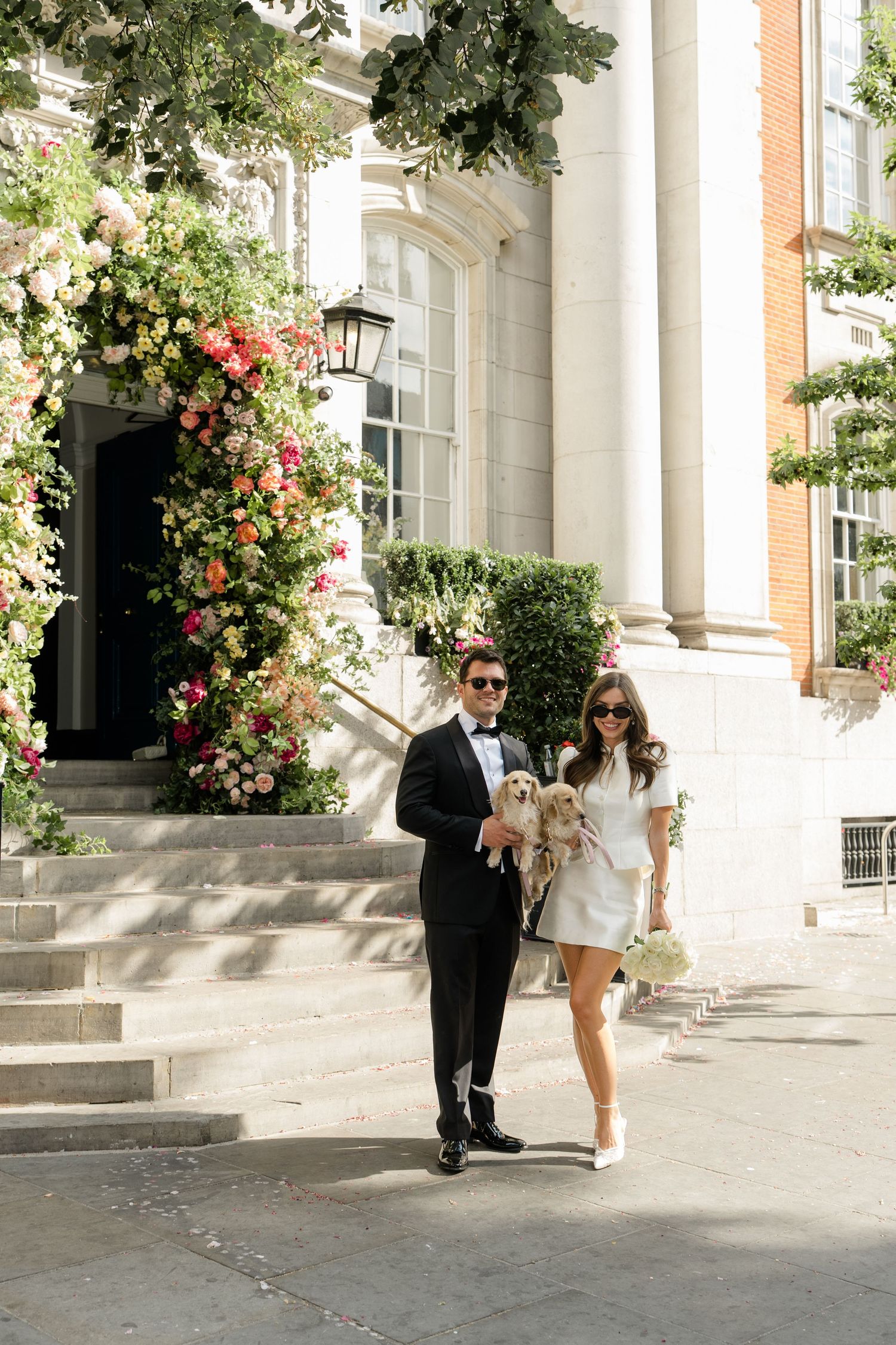 Couple in wedding attire holding a golden dog outside a floral-decorated elegant building entrance.