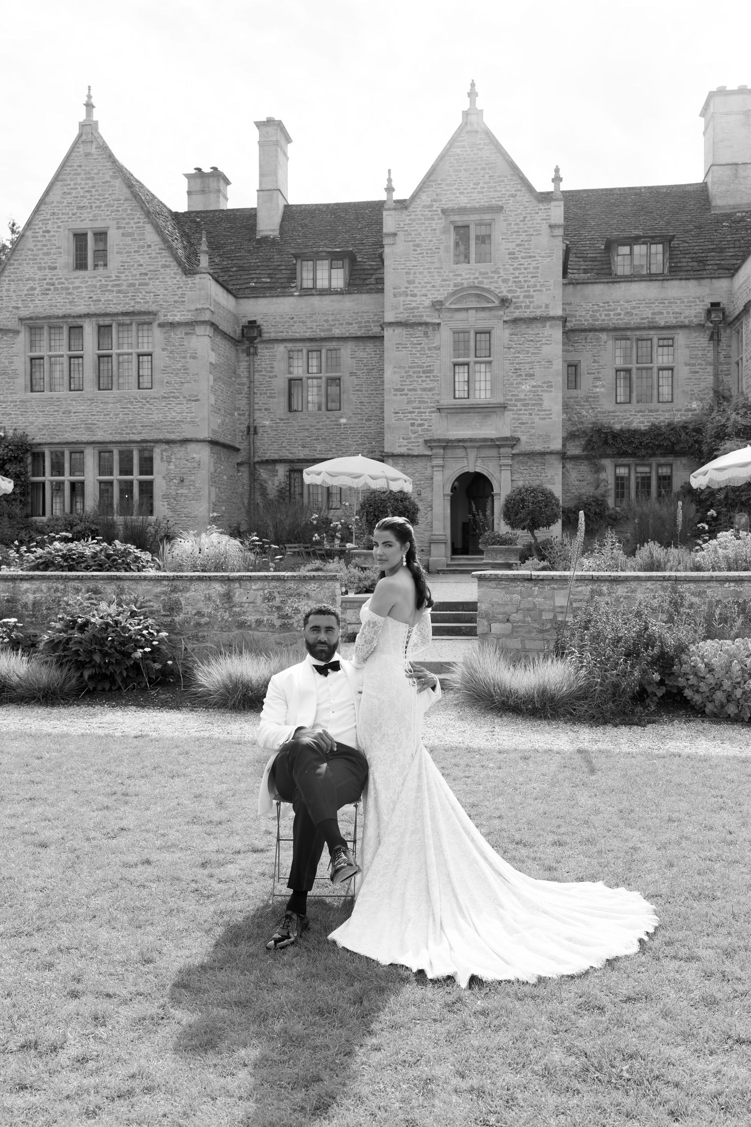 Black and white photo of bride and groom posing on lawn before grand historic English manor wedding venue.