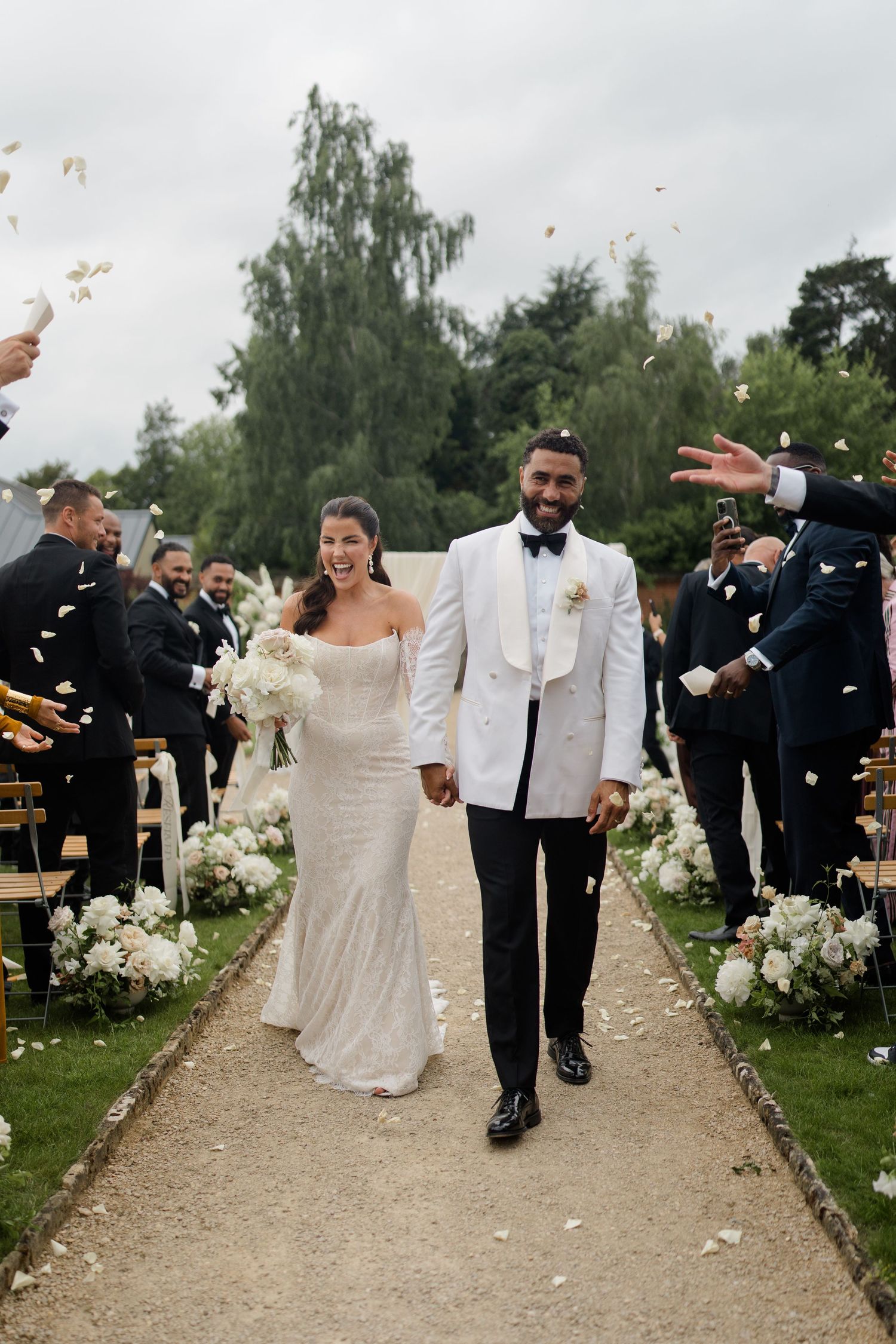 Bride and groom walk down outdoor aisle as guests throw white confetti at elegant garden wedding ceremony.