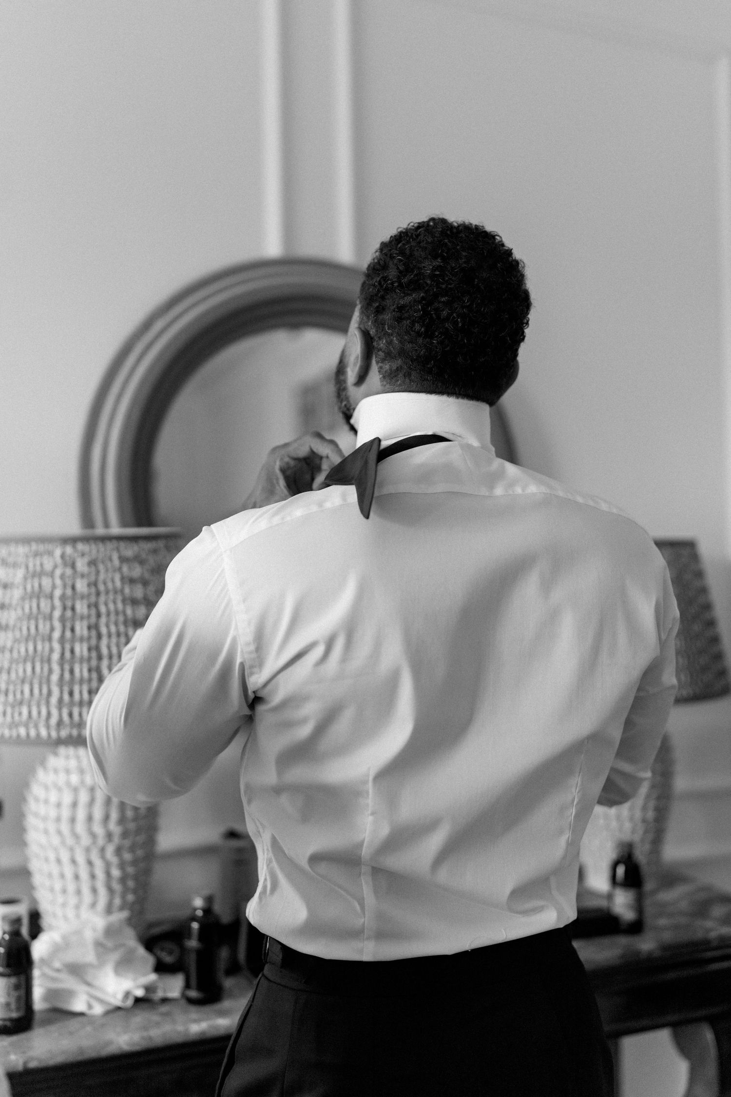 Groom in white dress shirt adjusting black bow tie while getting ready for wedding, rear view.