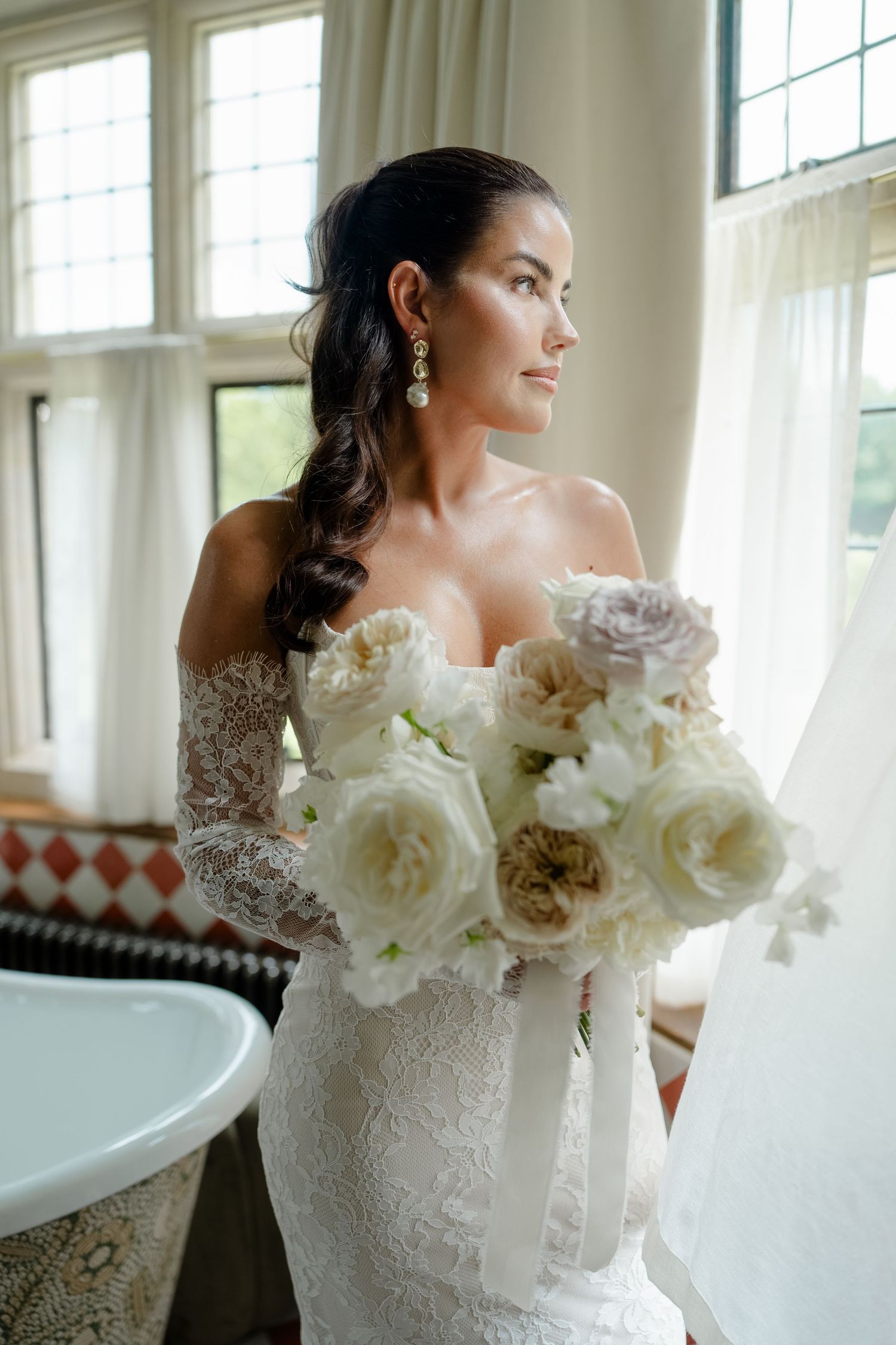 Bride in off-shoulder lace wedding gown holds white floral bouquet while gazing out window.