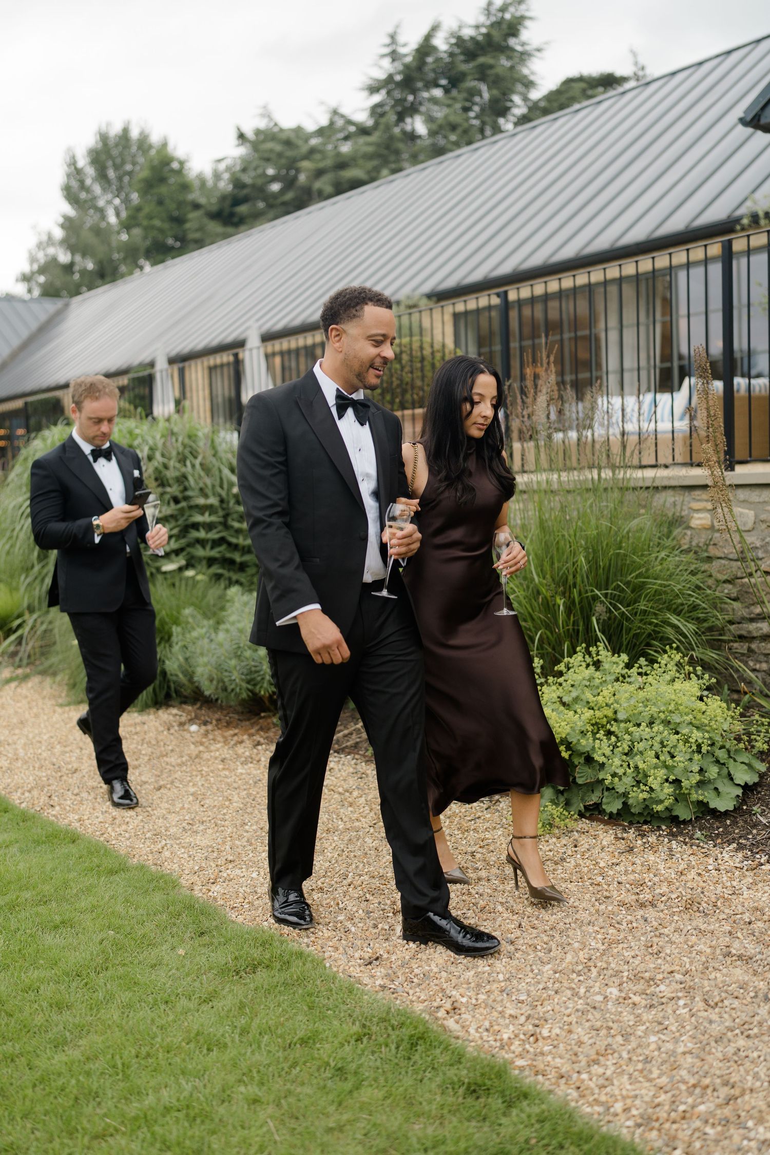 Wedding guests in formal attire walking along a gravel path outside a modern countryside venue.