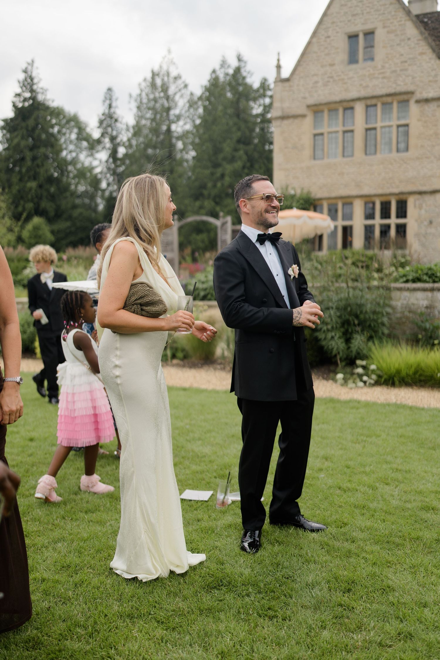 Wedding guests in formal attire laugh and socialize on manicured lawn outside elegant stone manor.
