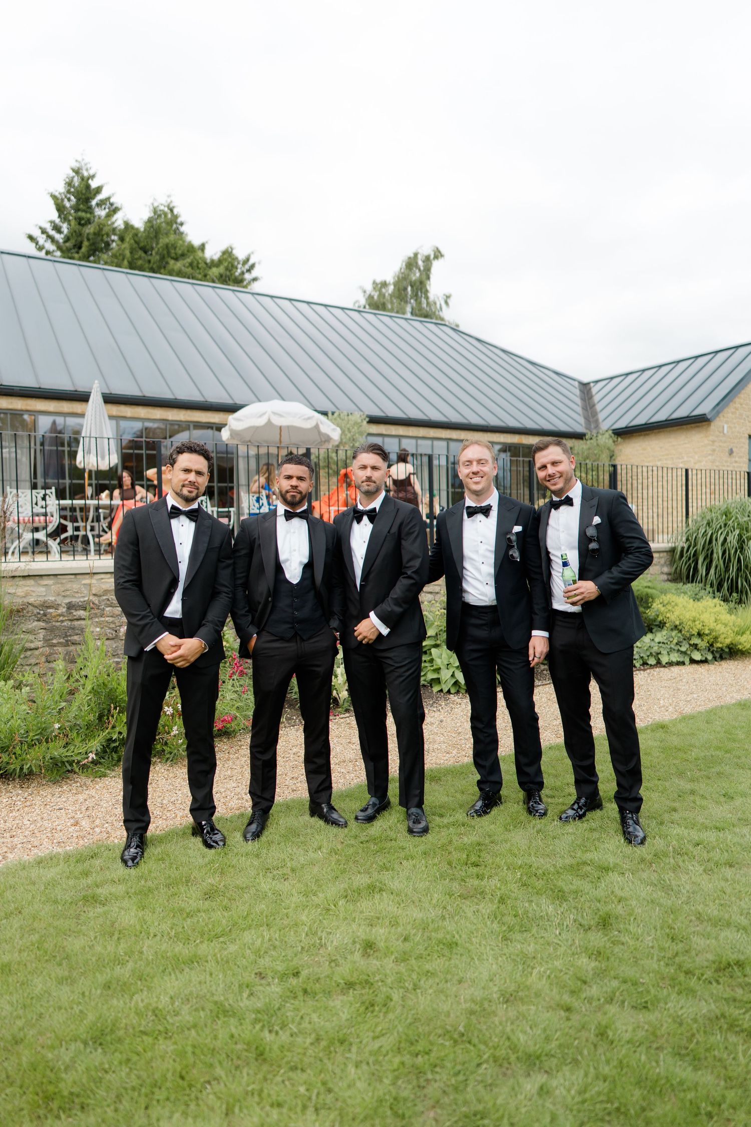 Five groomsmen in matching black tuxedos smiling together in a garden at a wedding reception.